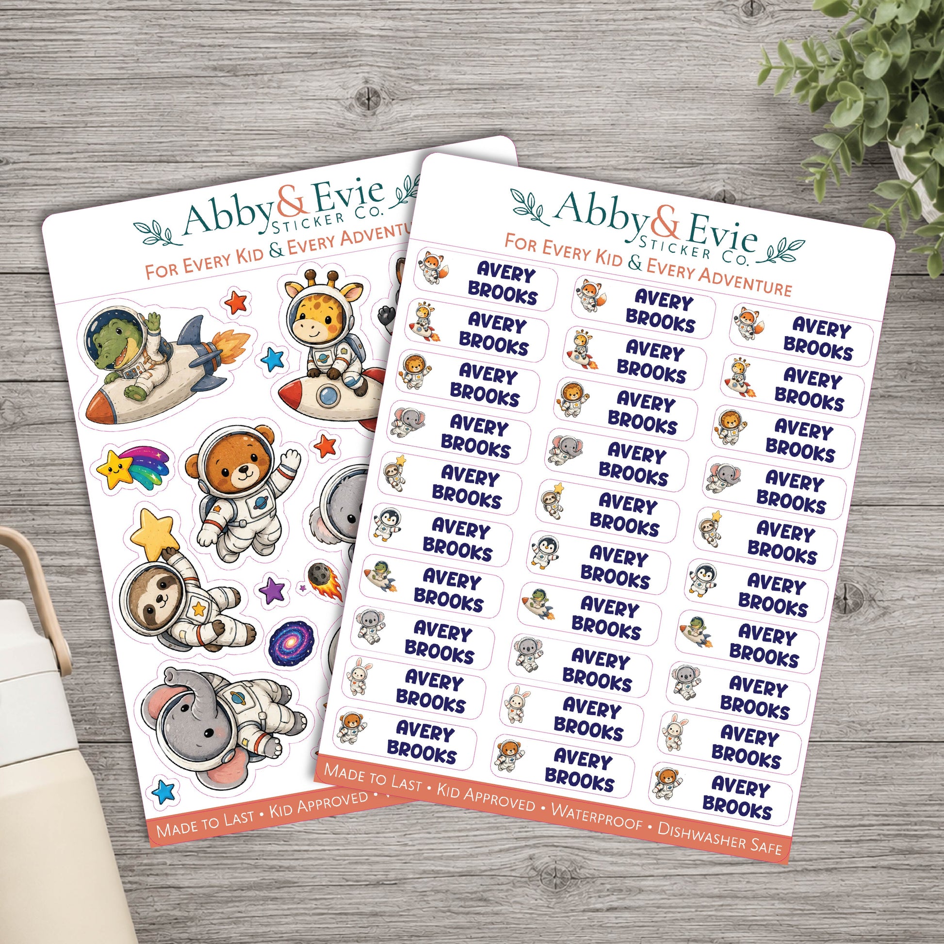 Two sticker sheets from Abby & Evie Sticker Co. featuring cartoon astronaut and space-themed characters, displayed on a gray wood background with a plant and notebook partially visible.