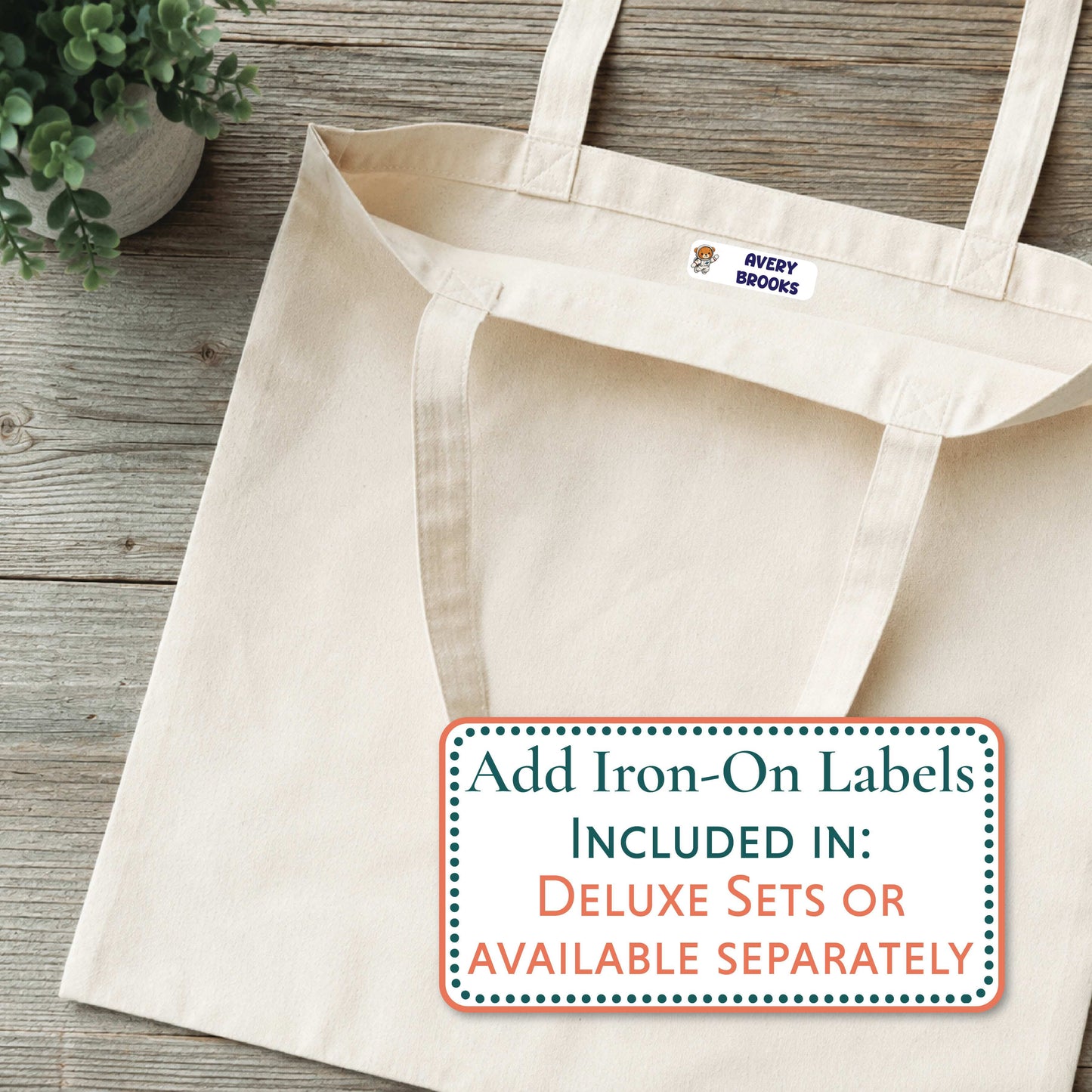 A natural canvas tote bag laid flat on a rustic wood surface, with a small purple Mabel logo patch near the top handle. A white promotional label overlay reads 'Add Iron-On Labels INCLUDED IN: DELUXE SETS OR AVAILABLE SEPARATELY.