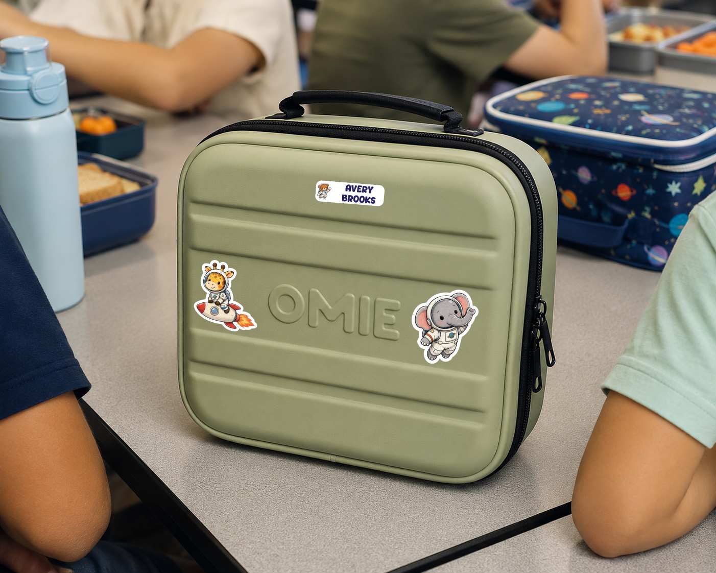 An olive green hard-shell lunch bag with a black handle and zipper, decorated with stickers and the name 'OMIE' on the front. It is shown on a classroom table surrounded by children and lunchboxes.