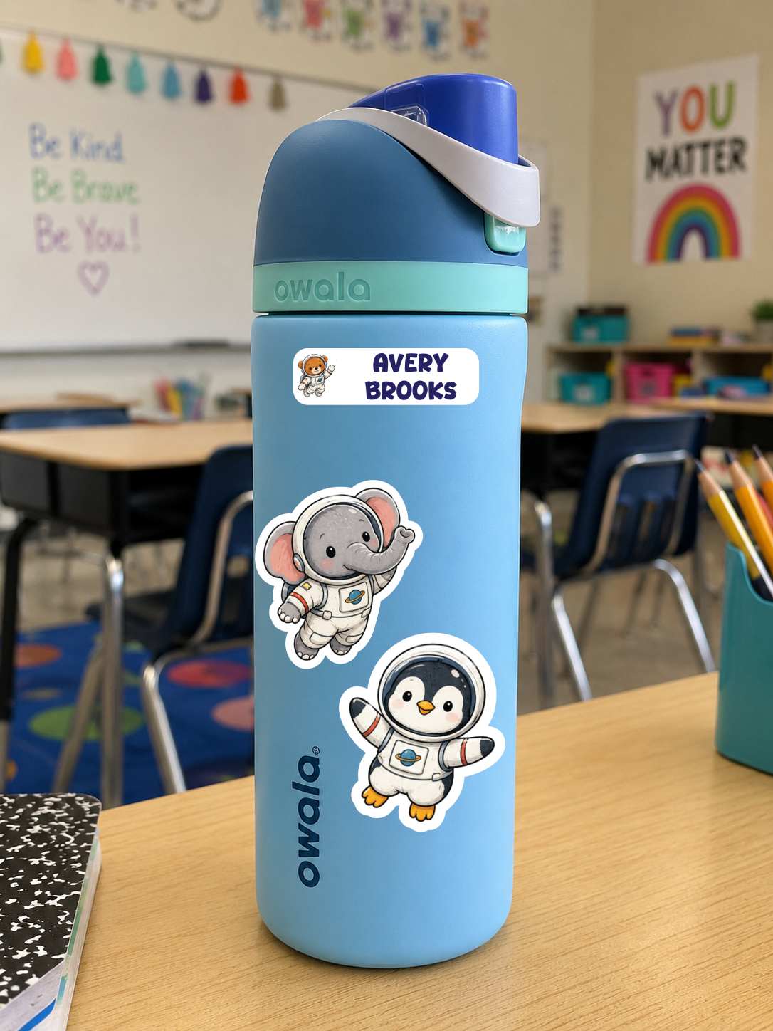 A light blue Owala water bottle with a dark blue lid and white strap, decorated with stickers of an elephant astronaut and a penguin astronaut, sitting on a wooden desk in a classroom.