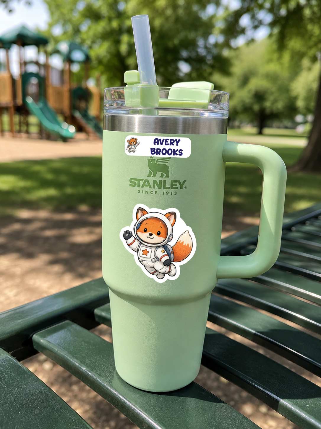 A mint green Stanley tumbler with a handle, clear lid, and straw, sitting on a green park bench. The tumbler features a sticker with the Avery Brooks Fox mascot and the Stanley logo.