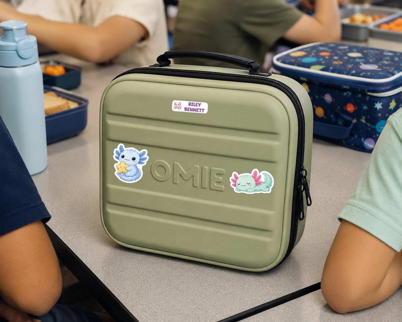 An olive green lunch bag with a black handle and zipper, decorated with stickers and the name 'OMIE', sitting on a school desk with children and lunchboxes in the background.
