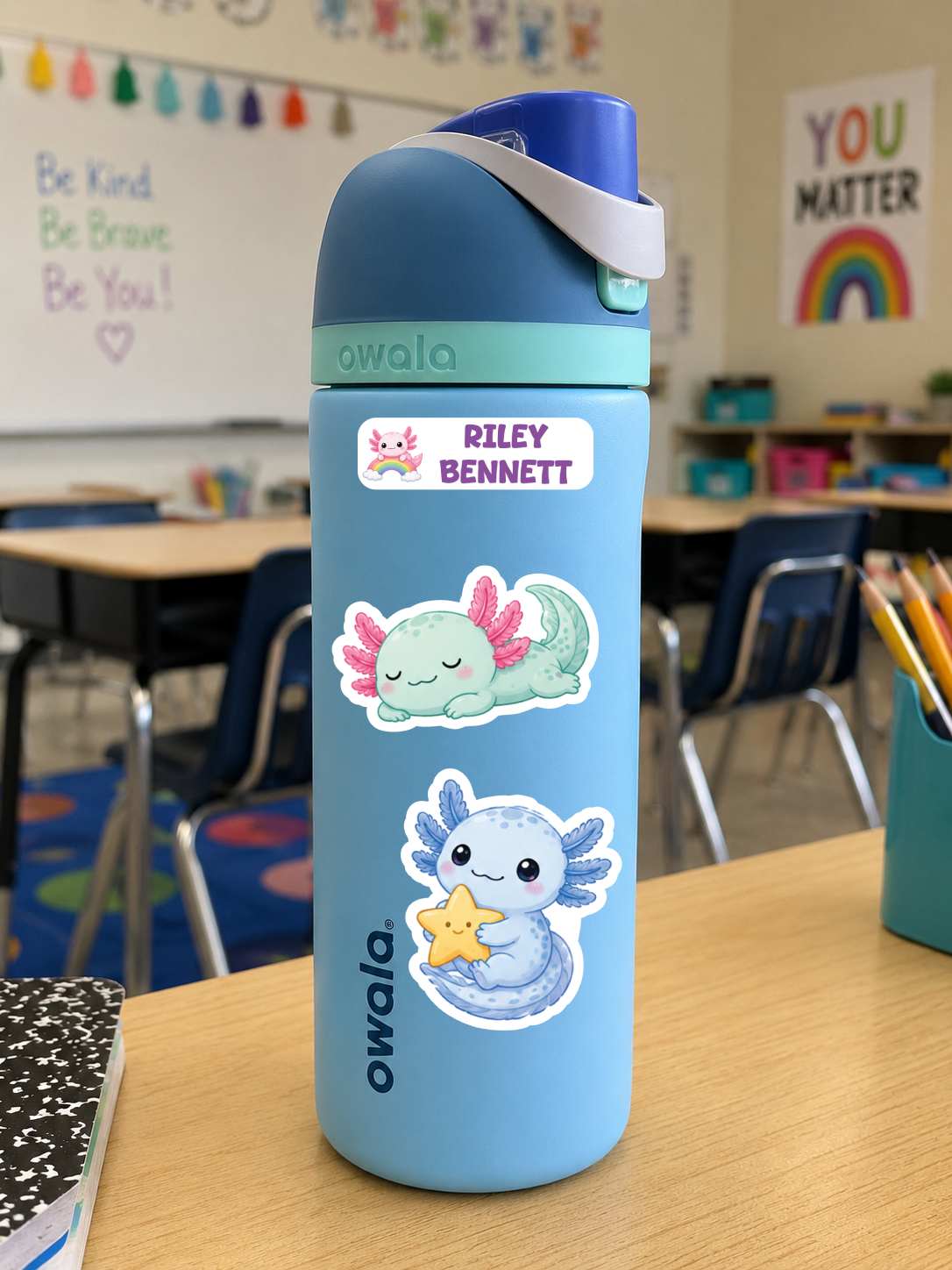 A light blue Owala water bottle with a custom name sticker and two axolotl stickers, standing on a wooden desk in a classroom.