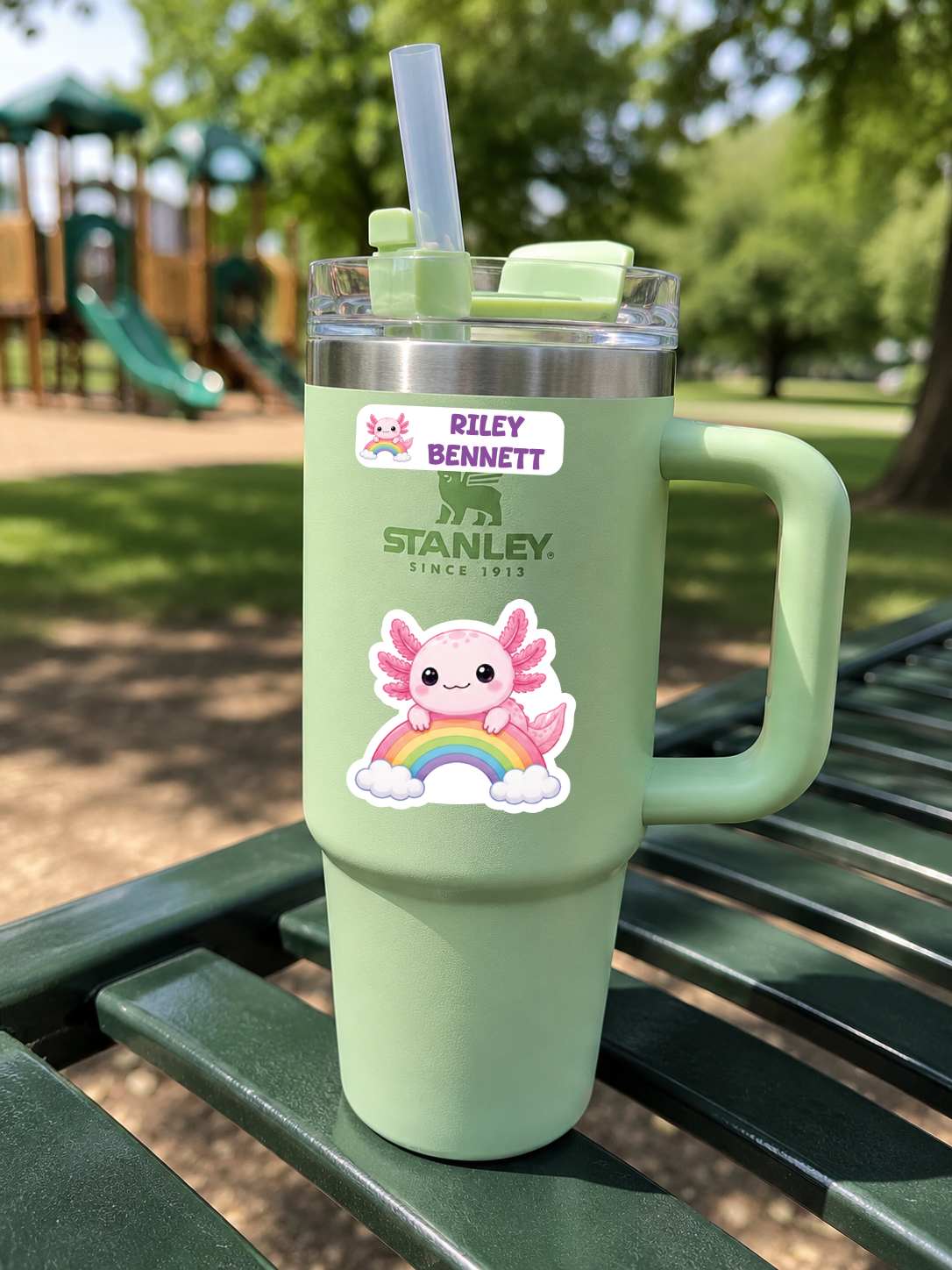 A mint green Stanley travel tumbler with a handle, clear lid, and straw, sitting on a green park bench. The tumbler has a custom sticker reading 'Riley Bennett' with a bear logo and a cute pink cartoon character with a rainbow.