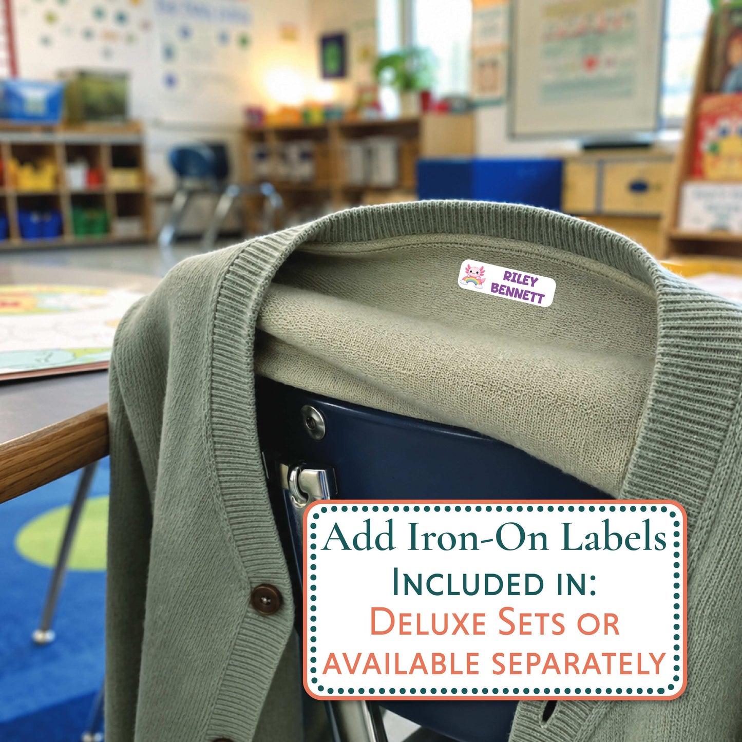An olive green knit cardigan sweater draped over a dark blue chair in a blurred classroom setting, with a small sewn-on label reading Riley Bennett visible inside the collar. A promotional text box at the bottom says add iron-on labels included in deluxe sets or available separately.