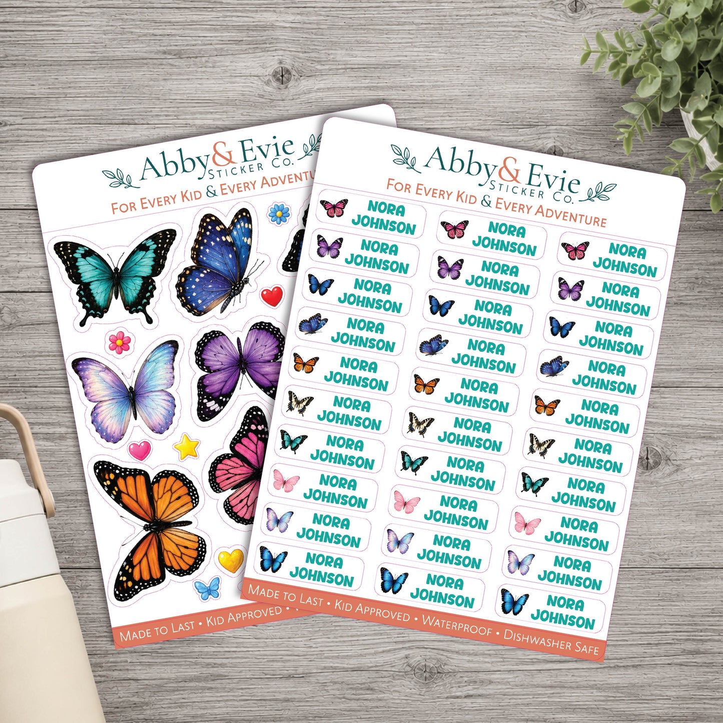 Two sticker sheets from Abby & Evie Sticker Co. featuring colorful butterfly illustrations and repeated 'Nora Johnson' names, arranged on a light wood surface with a small plant and book edge visible.