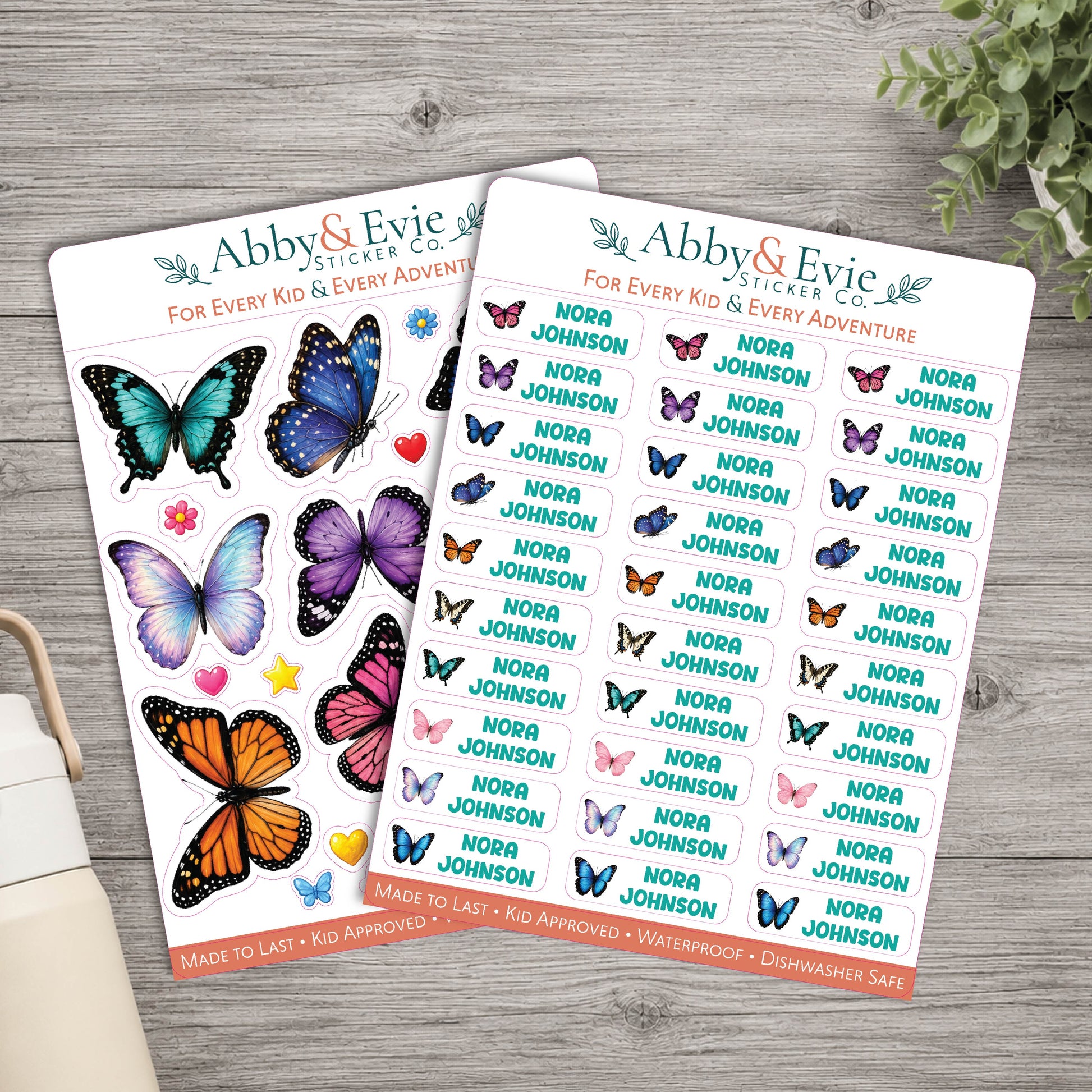 Two sticker sheets from Abby & Evie Sticker Co. featuring colorful butterfly illustrations and repeated 'Nora Johnson' names, arranged on a light wood surface with a small plant and book edge visible.