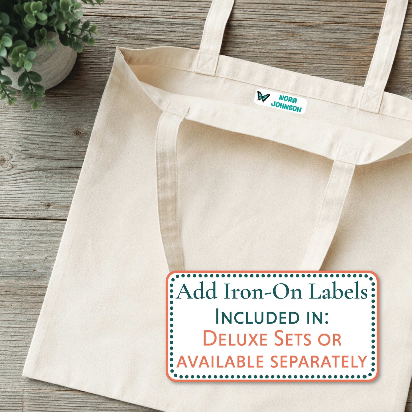 A natural canvas tote bag laid flat on a rustic wood surface, with a small blue and white 'GRAYSON' logo printed inside the neck. A large text overlay at the bottom right reads, 'Add Iron-On Labels INCLUDED IN: DELUXE SETS OR AVAILABLE SEPARATELY.