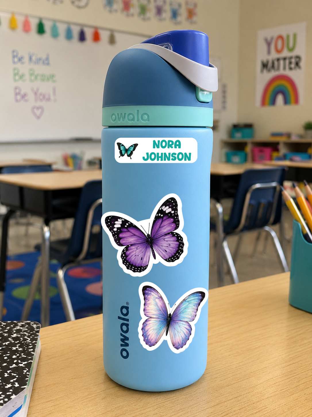 A light blue Owala water bottle with purple butterfly stickers and a custom name label reading 'NORA JOHNSON' sits on a wooden desk in a classroom setting.