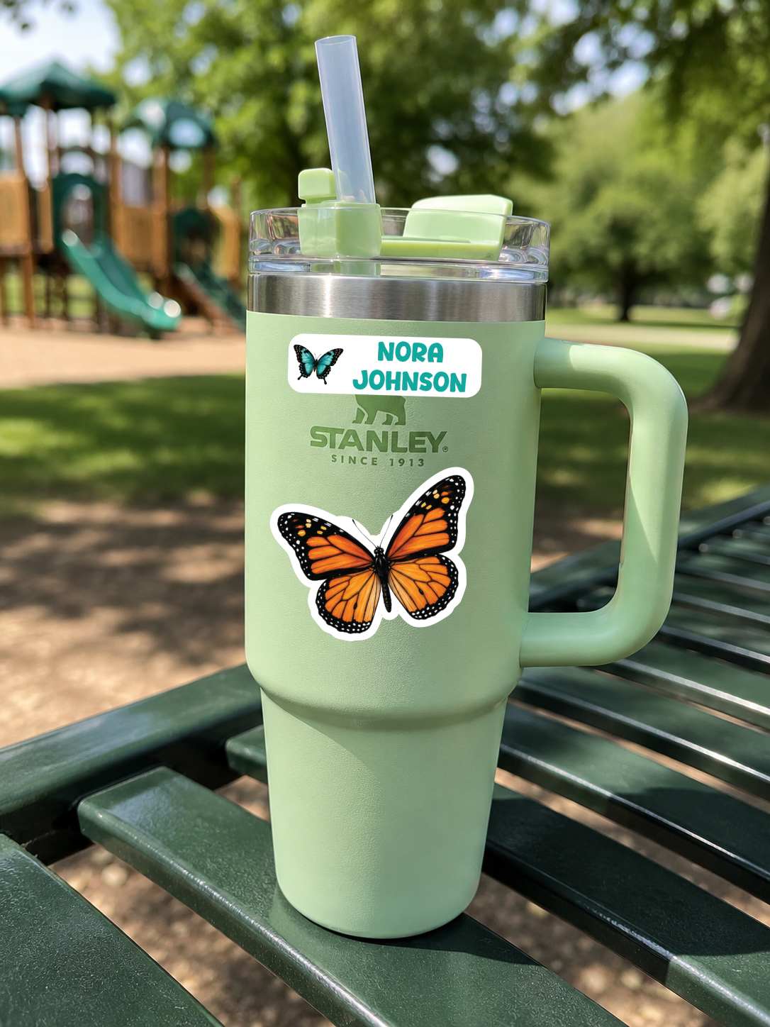 A mint green Stanley tumbler with a handle, clear lid, and straw sits on a green park bench. The tumbler has a custom name sticker reading 'NORA JOHNSON' and a large orange-and-black butterfly decal.