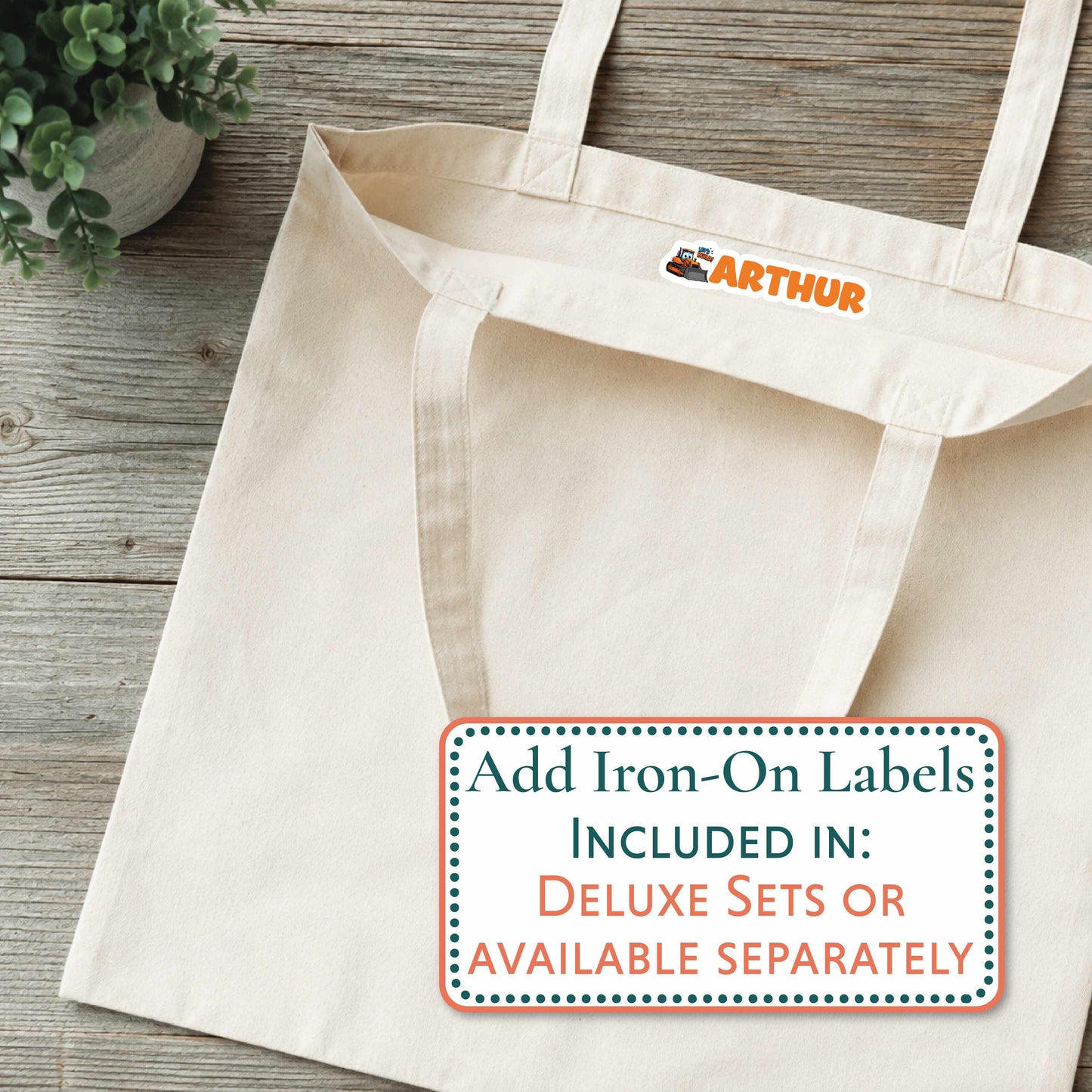 A cream canvas tote bag laid flat on a rustic wood surface, with a small orange 'ARTHUR' logo printed inside the neck area. A white promotional label at the bottom right reads 'Add Iron-On Labels INCLUDED IN: DELUXE SETS OR AVAILABLE SEPARATELY.