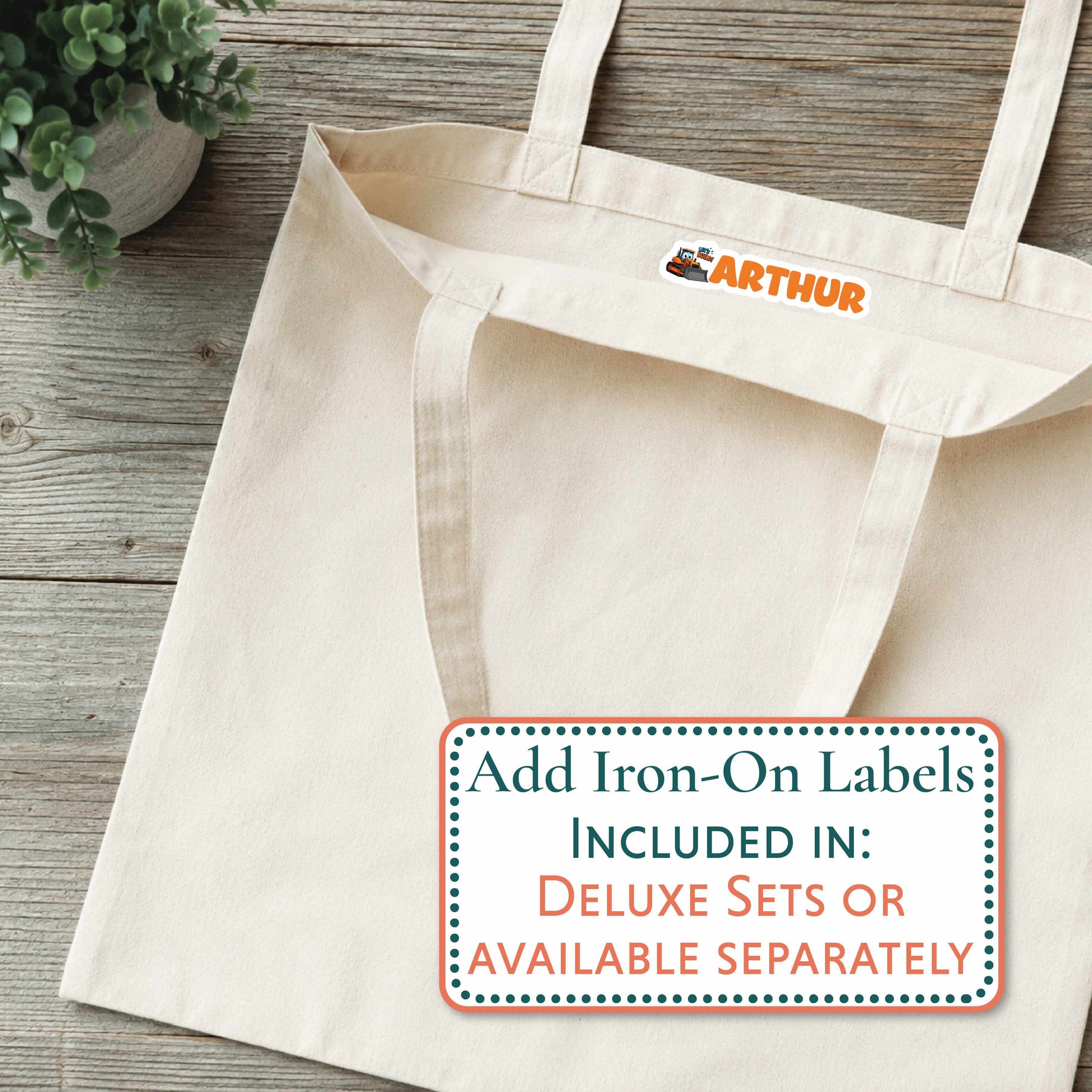 A cream canvas tote bag laid flat on a rustic wood surface, with a small orange 'ARTHUR' logo printed inside the neck area. A white promotional label at the bottom right reads 'Add Iron-On Labels INCLUDED IN: DELUXE SETS OR AVAILABLE SEPARATELY.