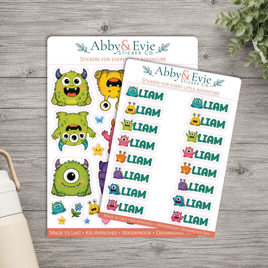 Two sheets of Abby & Evie sticker co. character stickers on a gray wood background, with a small portion of a white bag and green leaves visible at the edges.