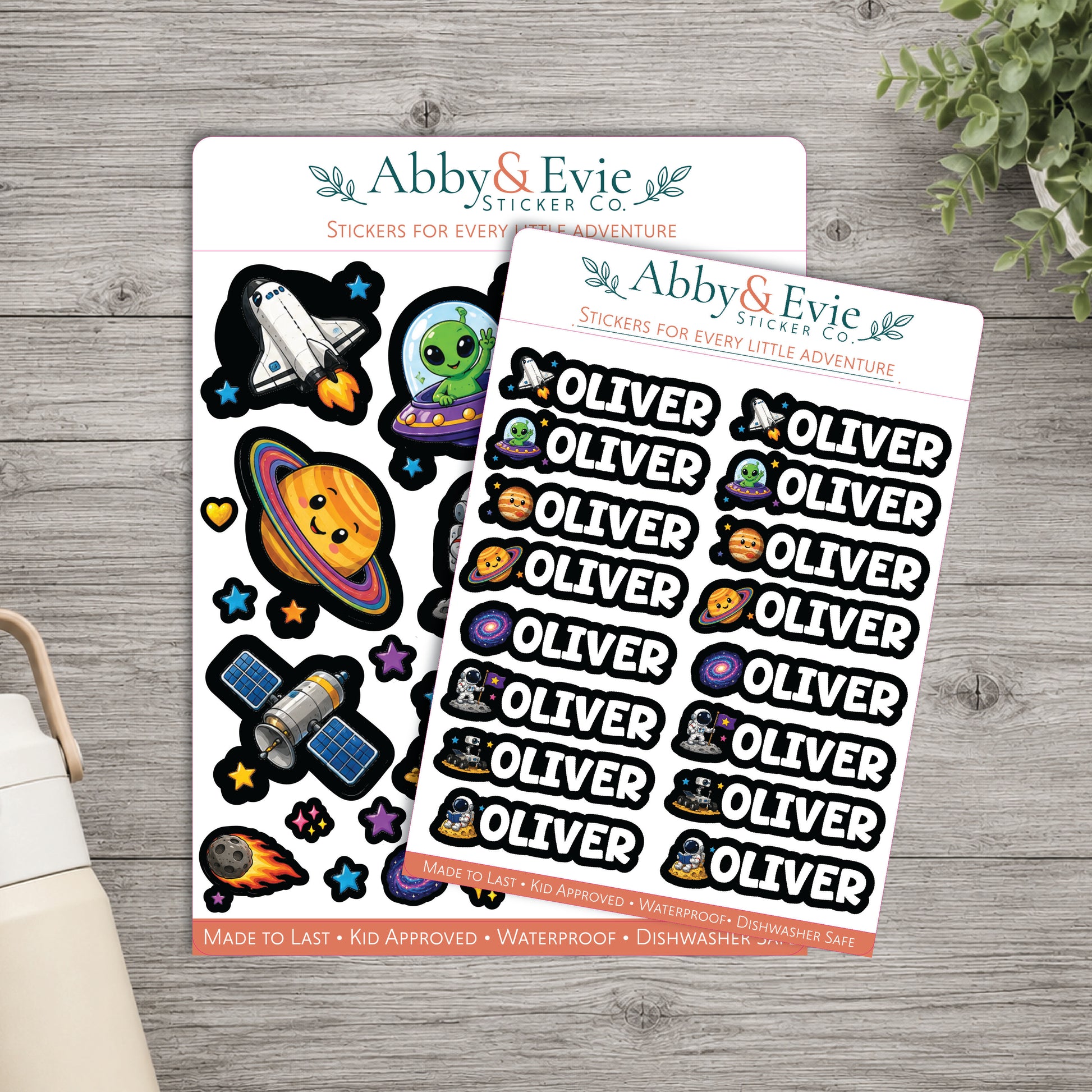 Two sheets of Abby & Evie stickers on a gray wood tabletop, with one sheet featuring space-themed stickers and the other featuring the name Oliver repeated across the sheet. A small plant and part of a bag are visible at the edges.