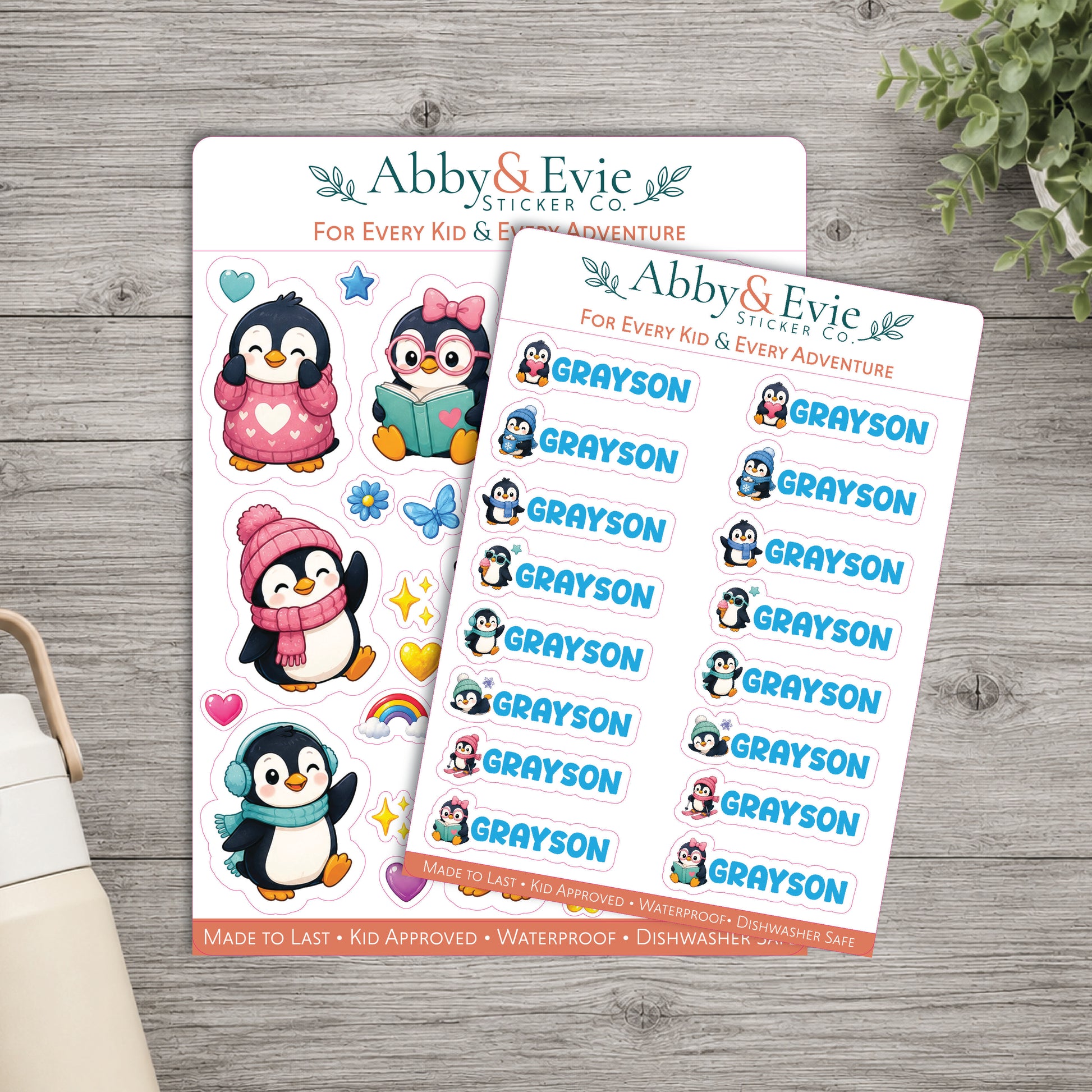 Two sticker sheets from Abby & Evie Sticker Co. featuring cartoon penguins and the name 'Grayson' on a gray wood background, with a small plant and part of a bag visible at the edges.