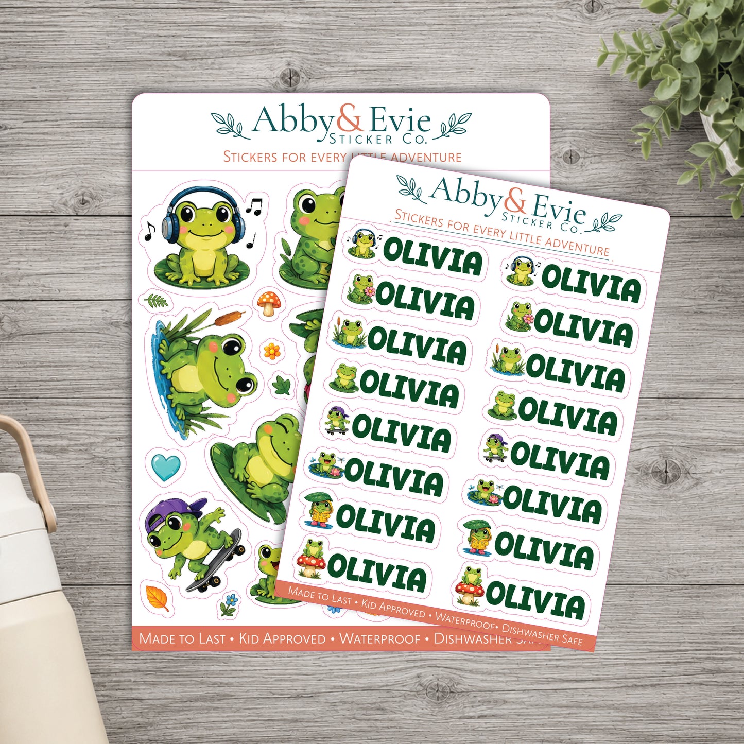 Two sheets of cartoon frog stickers from Abby & Evie Sticker Co are displayed on a gray wood background, with a small plant leaf and part of a mug visible at the edges.