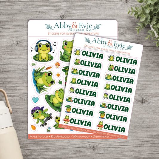 Two sheets of cartoon frog stickers from Abby & Evie Sticker Co are displayed on a gray wood background, with a small plant leaf and part of a mug visible at the edges.