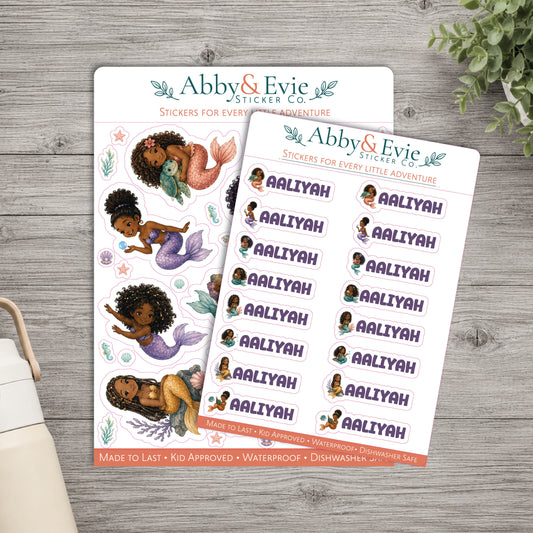 Two sticker sheets from Abby & Evie Sticker Co. featuring mermaid illustrations and the name 'Aaliyah' repeated in purple text, arranged on a gray wood surface with a small plant and part of a bag visible.