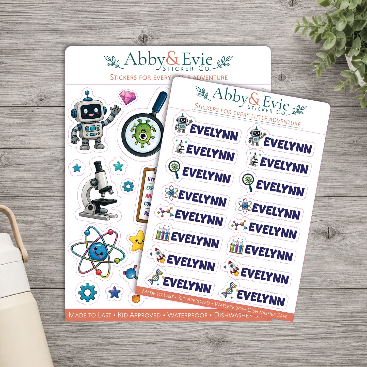 Two sheets of Abby & Evie sticker set stickers for 'Every Little Adventure' displayed on a gray wood background with a plant and part of a mug visible.