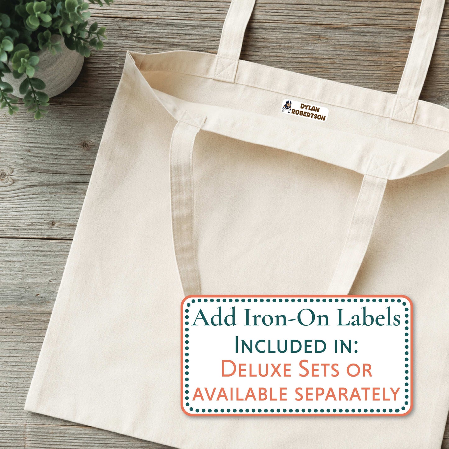 A natural canvas tote bag laid flat on a rustic wood surface, with a small blue and white 'GRAYSON' logo printed inside the neck. A large text overlay at the bottom right reads, 'Add Iron-On Labels INCLUDED IN: DELUXE SETS OR AVAILABLE SEPARATELY.