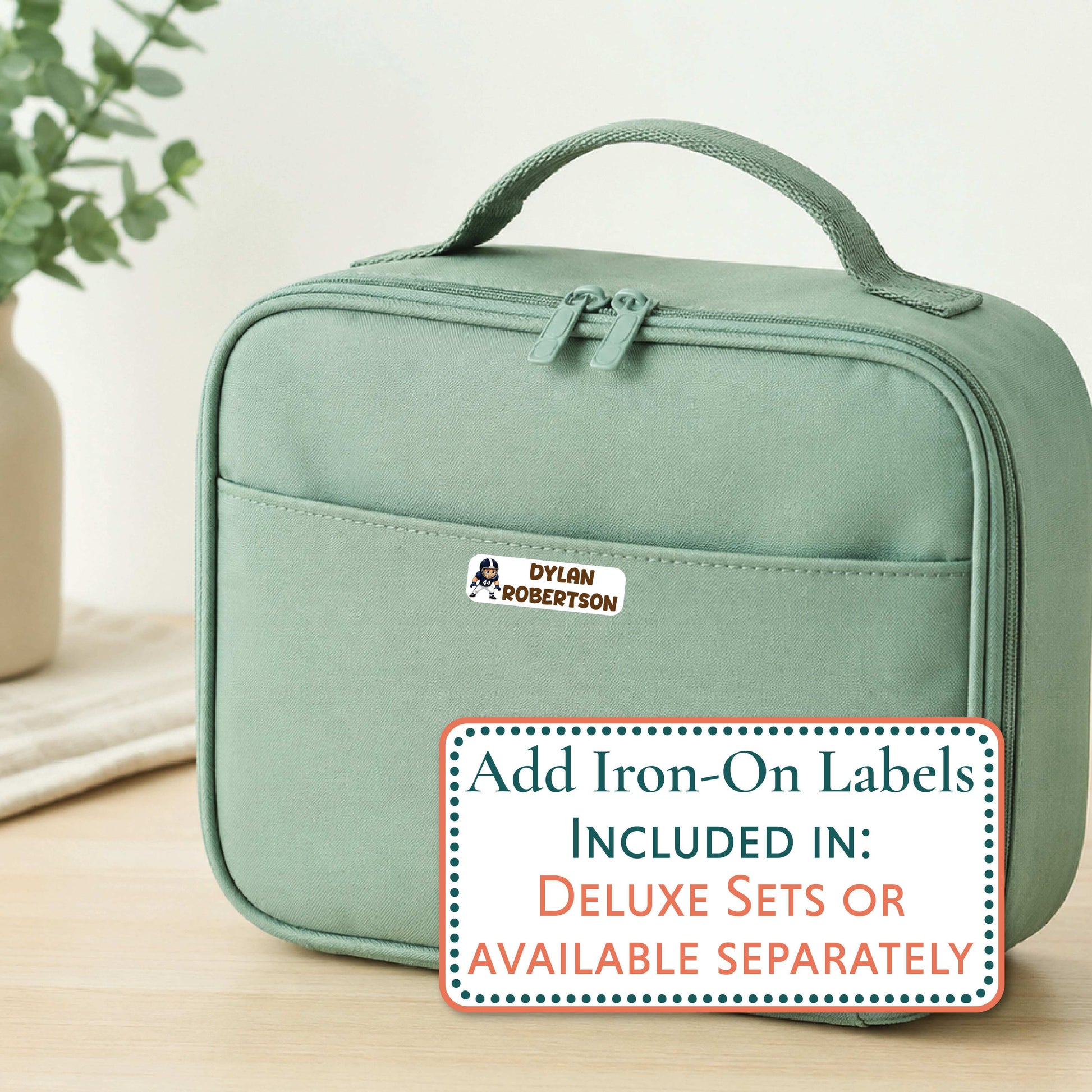 A sage green insulated lunch bag with a top handle and front zip pocket, shown on a light wood surface with a decorative plant in the background. A white sticker with the name 'Aaliyah' is visible on the front pocket, and a promotional text overlay at the bottom reads 'Add Iron-On Labels INCLUDED IN: DELUXE SETS OR AVAILABLE SEPARATELY'.