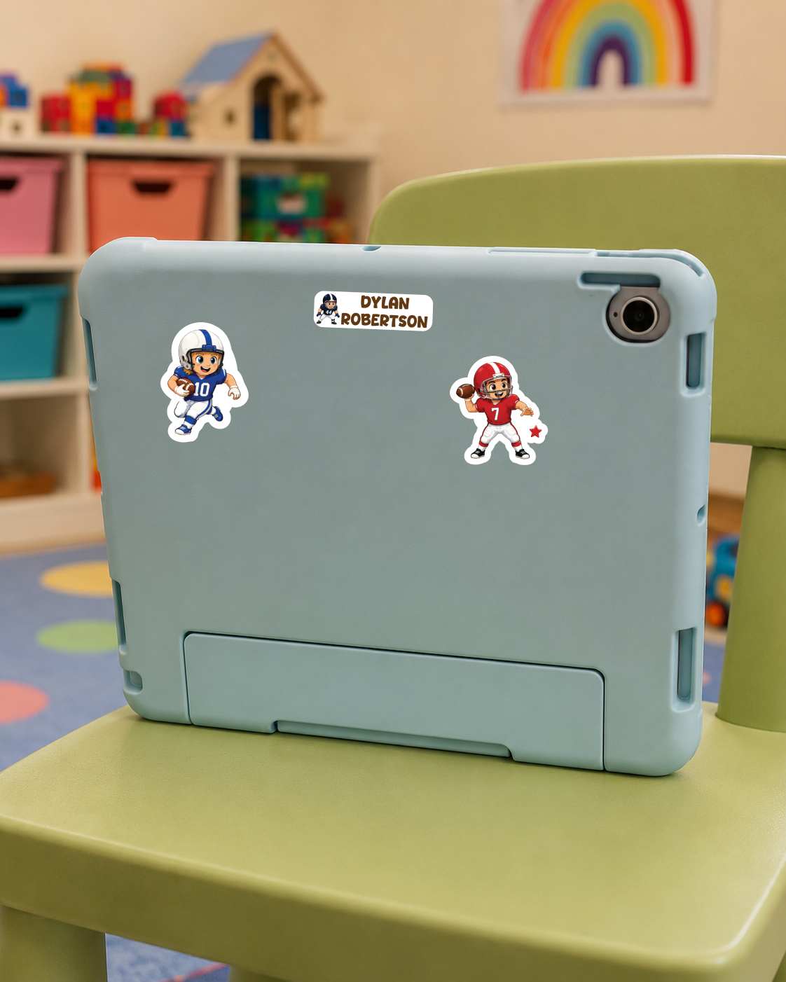 A light blue tablet case with two football-themed stickers is shown on a green chair in a child's room. The stickers feature cartoon characters and the name 'Dylan Robertson'.