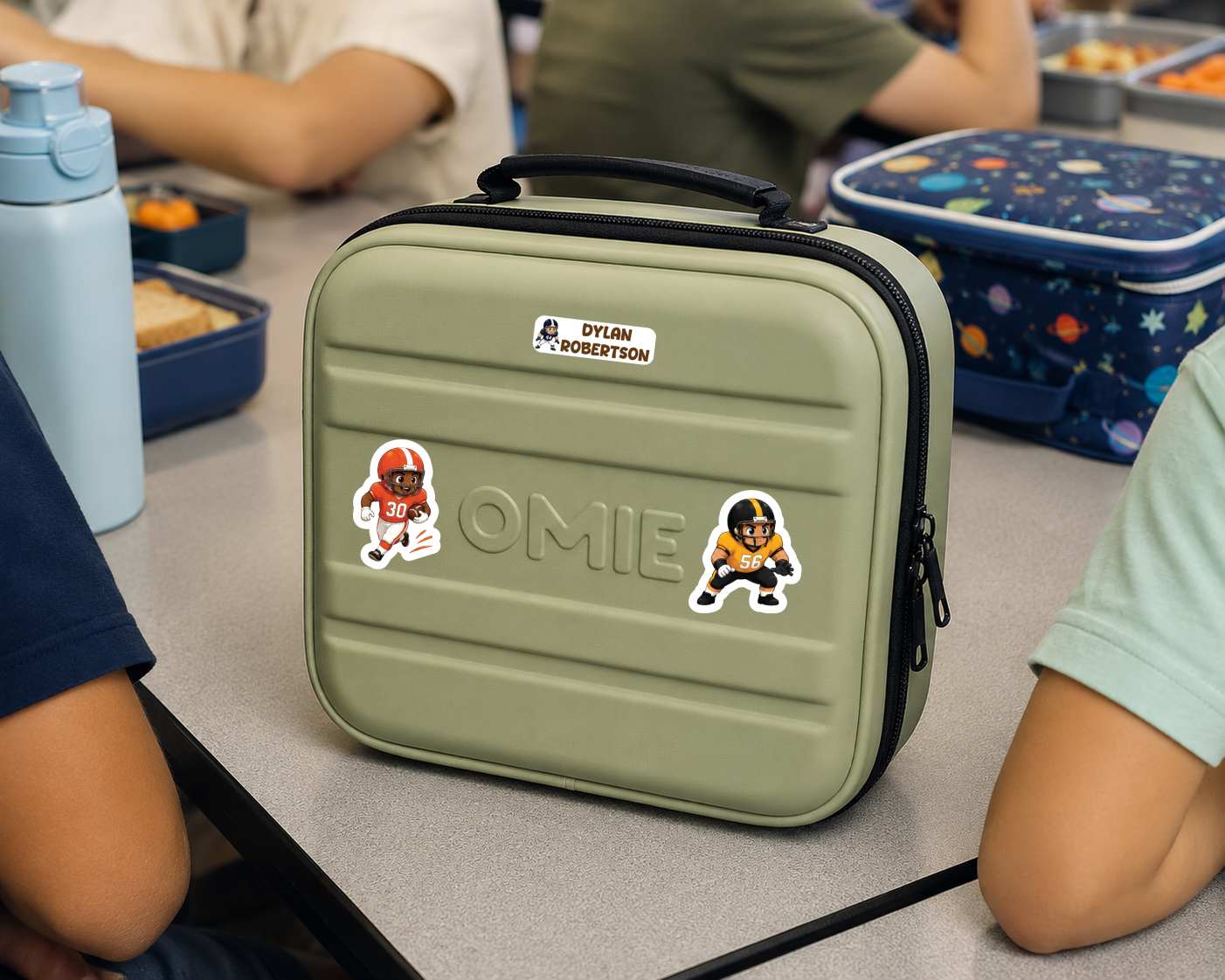 A customized olive green hard-sided lunch bag with black zippers and a top handle, decorated with a 'Dylan Roberts' name sticker and two football player stickers. The bag is shown on a school desk surrounded by children and other lunchboxes.