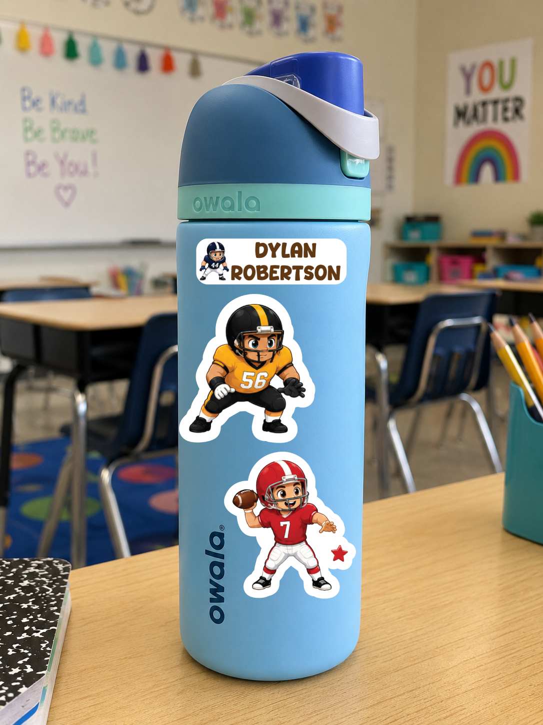 A light blue Owala water bottle with a dark blue lid and white strap, decorated with two football player stickers, standing on a wooden desk in a classroom.