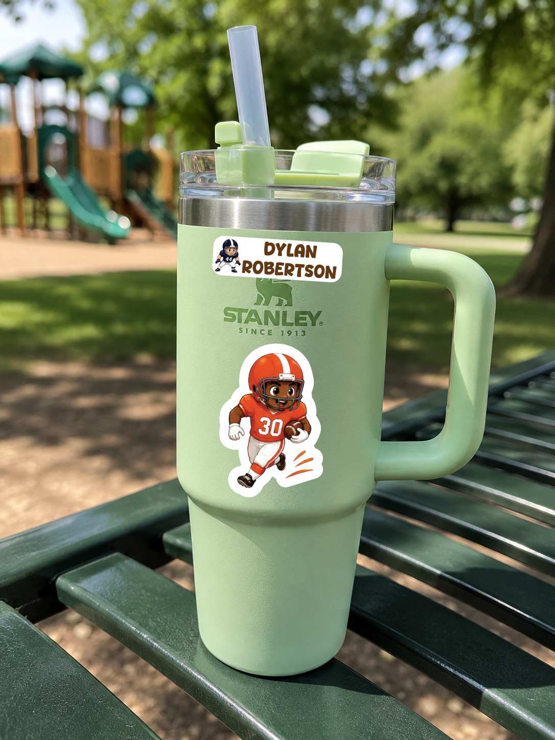 A mint green Stanley tumbler with a handle, clear lid, and straw, placed on a green park bench outdoors. The tumbler has a sticker reading 'Dylan Robertson' and another football player sticker.
