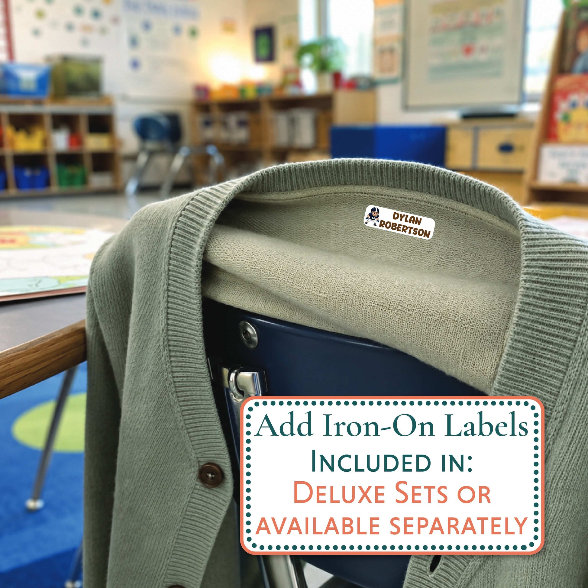 An olive green knit cardigan sweater draped over a dark chair in a blurred classroom setting, with a small pink and white 'CHARLOTTE' iron-on label visible inside the collar. A text overlay at the bottom reads, 'Add Iron-On Labels Included in: Deluxe Sets or Available Separately.