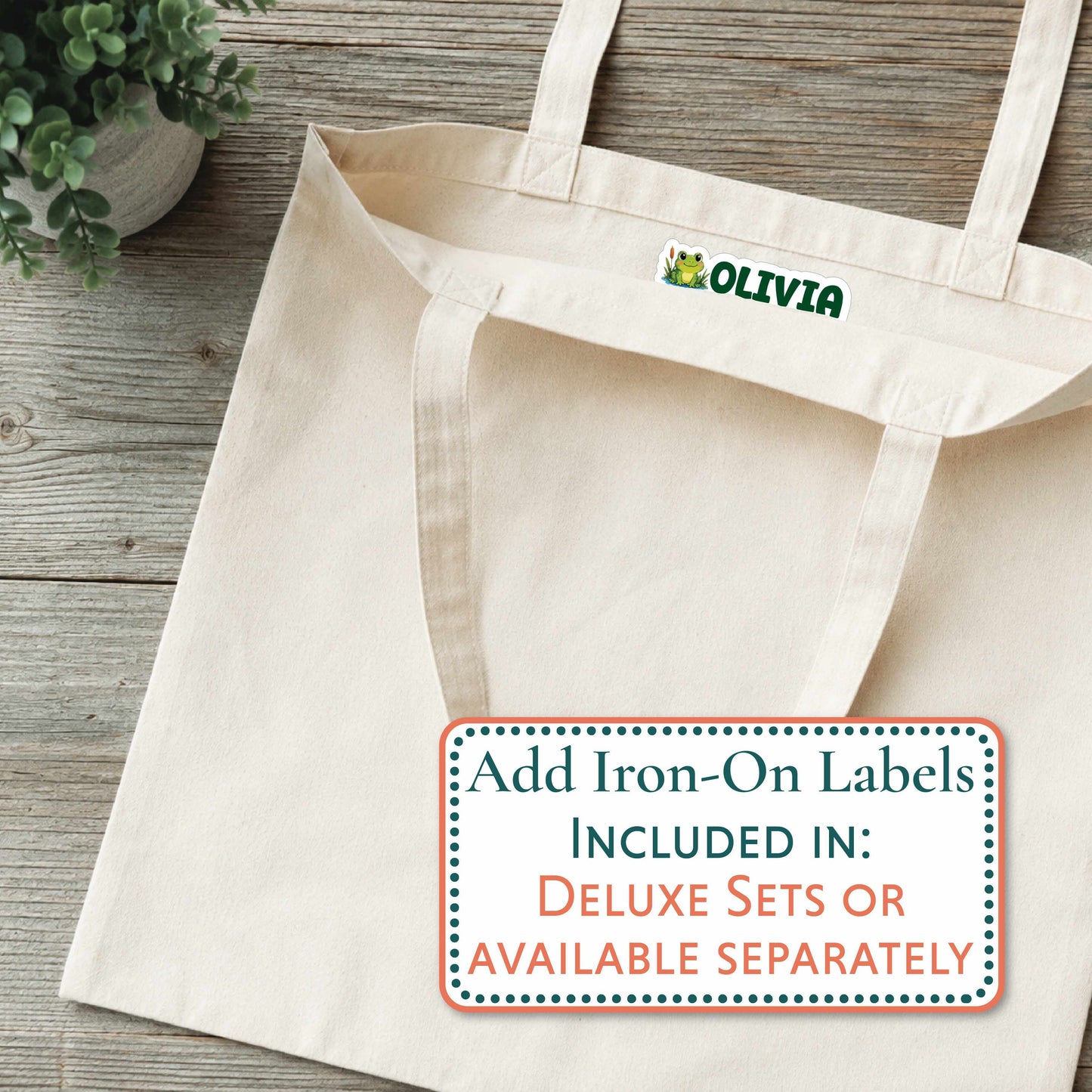 A cream canvas tote bag laid flat on a rustic wood surface, with a small green and yellow frog logo and the word OLIVIA printed inside the neck. A rectangular text overlay at the bottom right says, 'Add Iron-On Labels INCLUDED IN: DELUXE SETS OR AVAILABLE SEPARATELY.