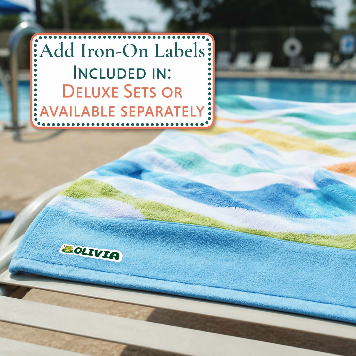 A colorful beach towel with a blue border and a small OLIVIA logo is draped over a poolside lounge chair, with a swimming pool and outdoor patio in the background. A text box in the upper left reads, 'Add Iron-On Labels INCLUDED IN: DELUXE SETS OR AVAILABLE SEPARATELY'.