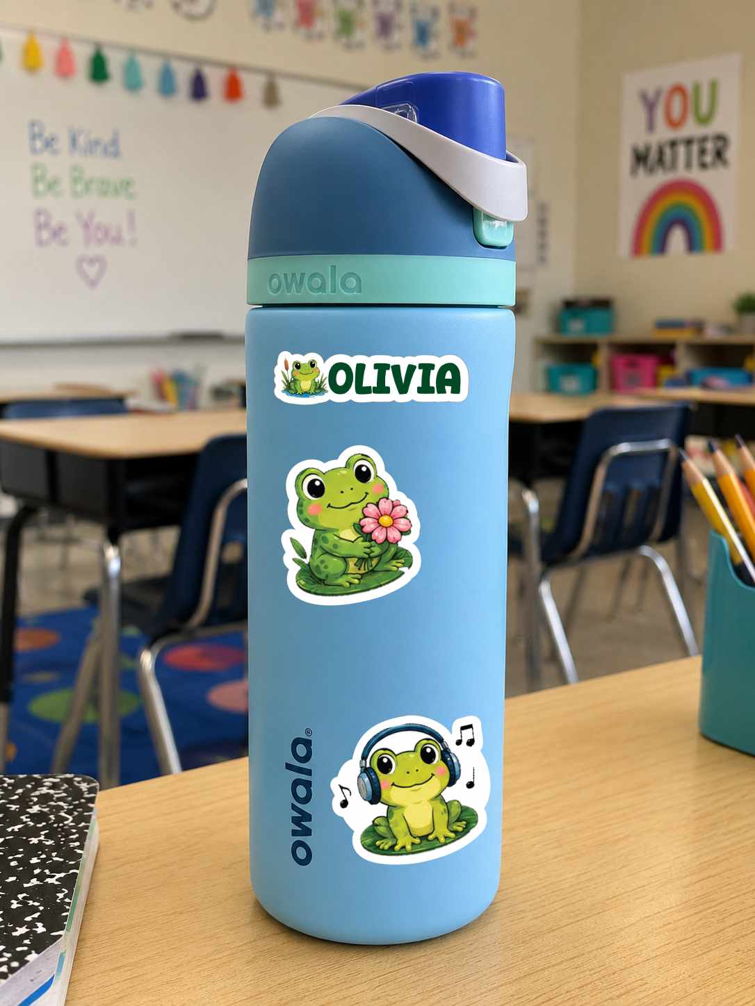 A light blue Owala water bottle with a dark blue lid and white strap, decorated with two frog stickers and the name 'OLIVIA' on a classroom desk.