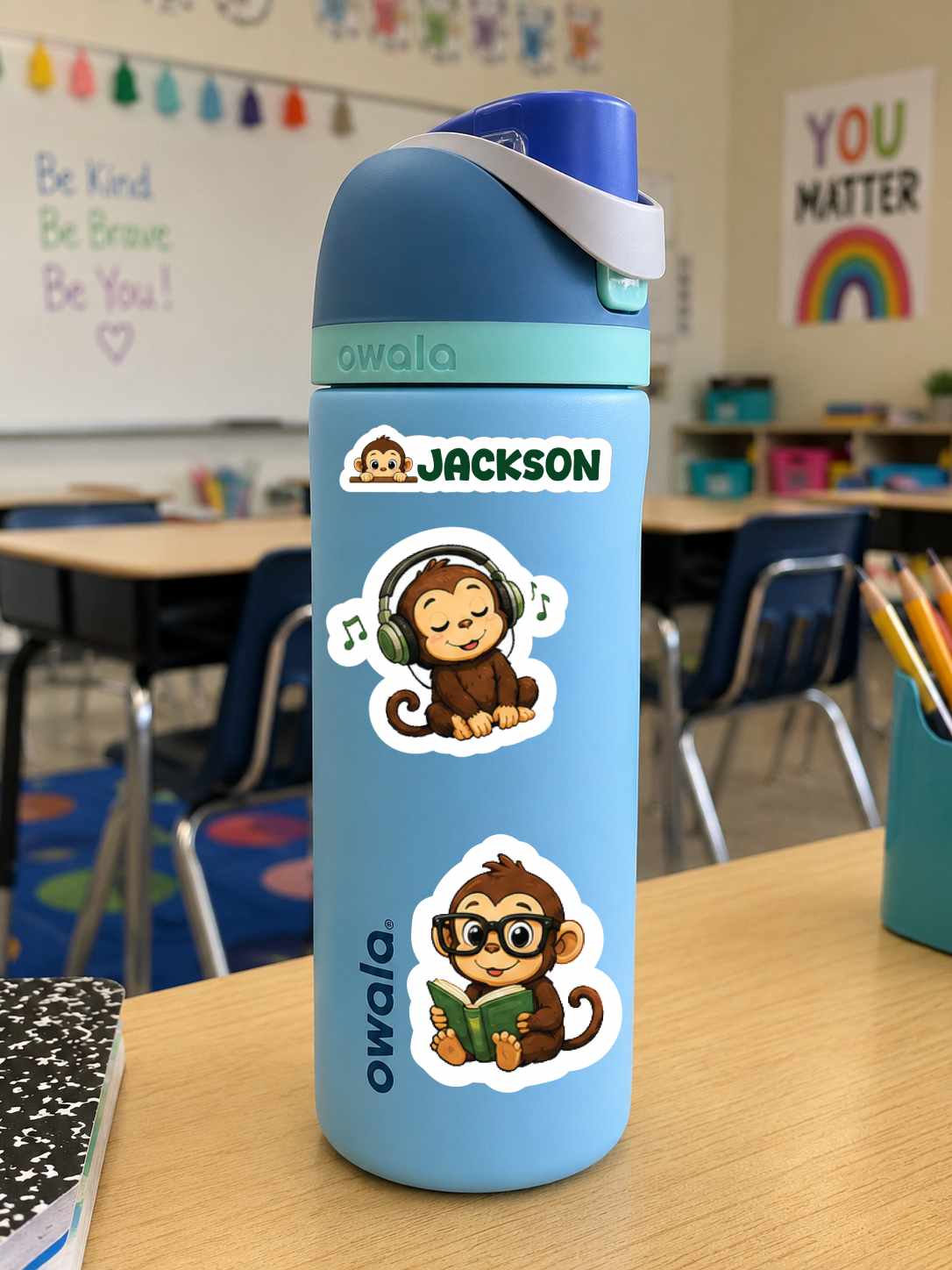 A light blue Owala water bottle with a custom name sticker and cartoon monkey decals, sitting on a wooden desk in a classroom setting.