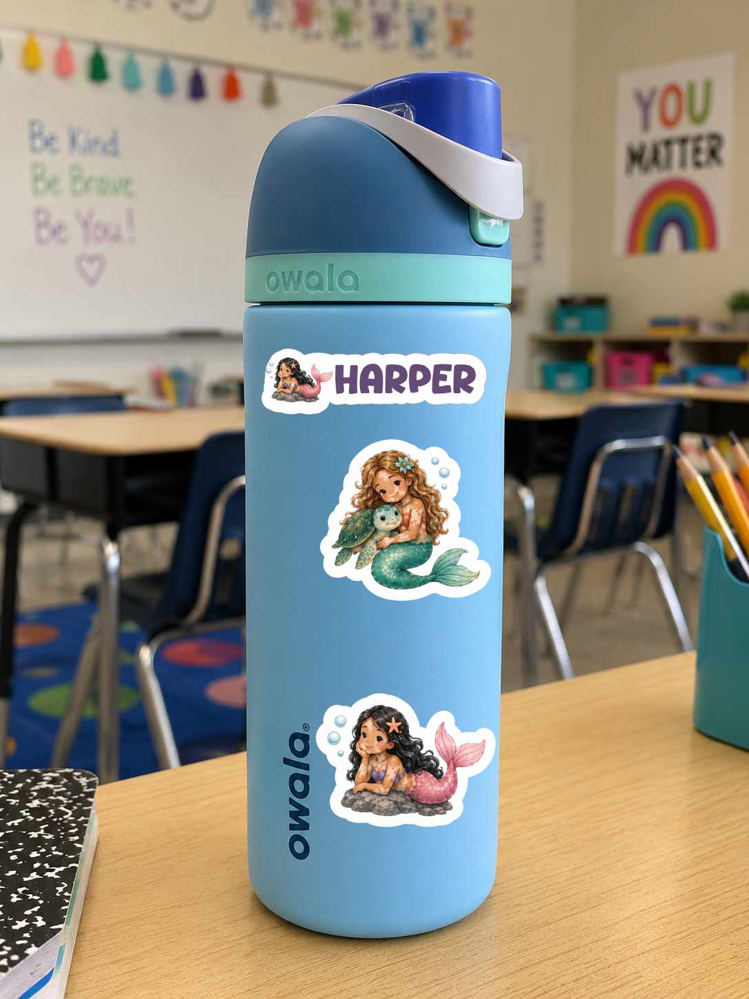 A light blue Owala water bottle with a dark blue lid and white strap, decorated with three mermaids with vitiligo stickers, standing on a wooden desk in a classroom.