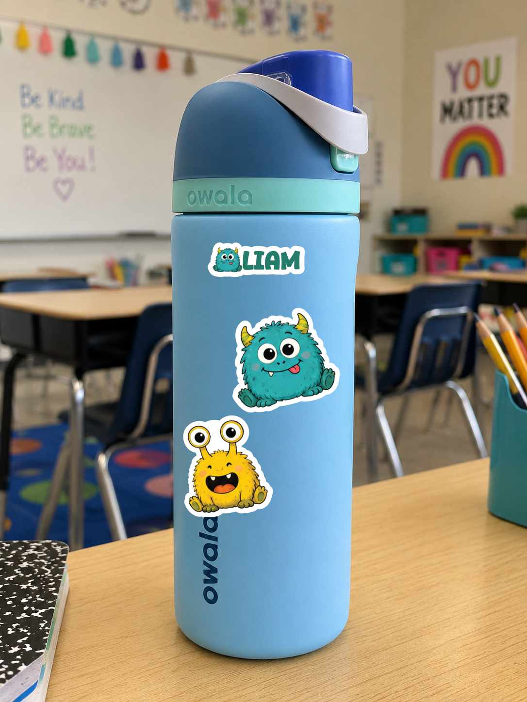 A light blue Owala water bottle with a dark blue lid and white strap, decorated with two monster stickers, sitting on a wooden desk in a classroom.