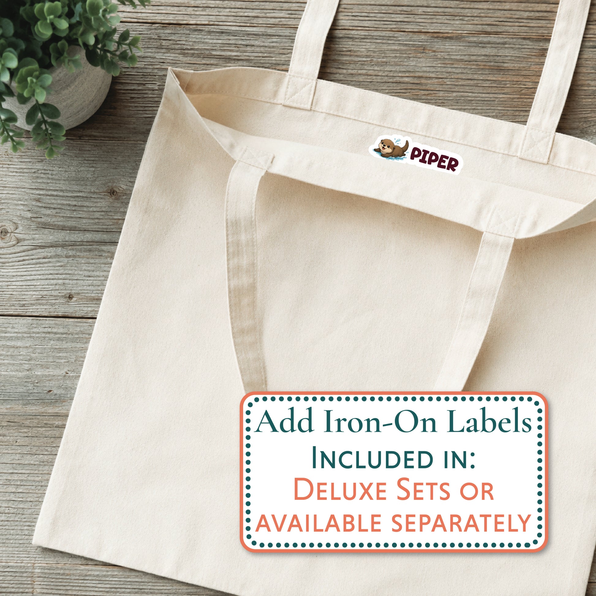 A natural canvas tote bag laid flat on a rustic wood surface, with a small embroidered 'PIPER' logo near the top. A large text label in the lower right says 'Add Iron-On Labels INCLUDED IN: DELUXE SETS OR AVAILABLE SEPARATELY.