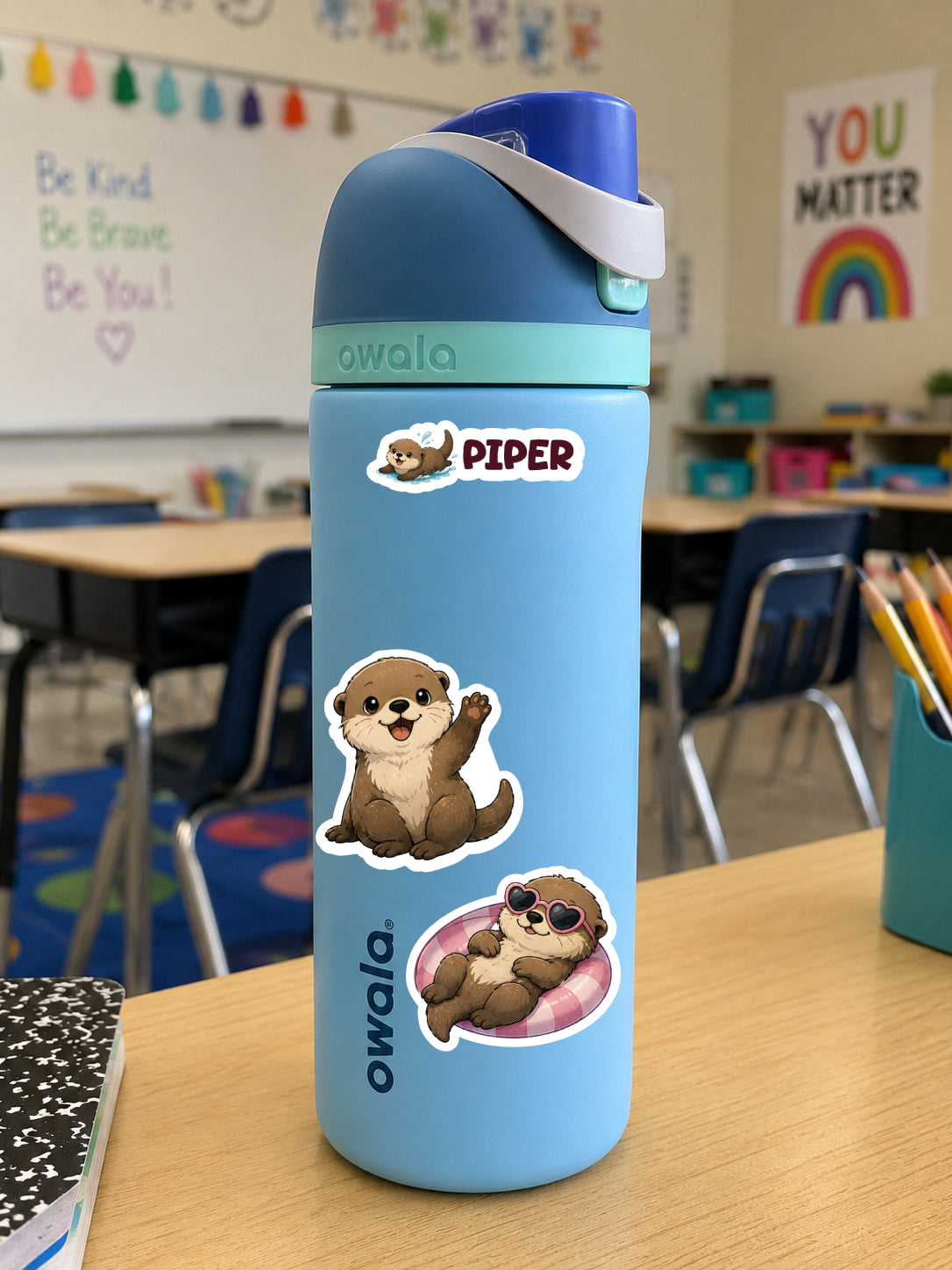 A light blue Owala water bottle with cartoon otter stickers, placed on a wooden desk in a classroom setting.