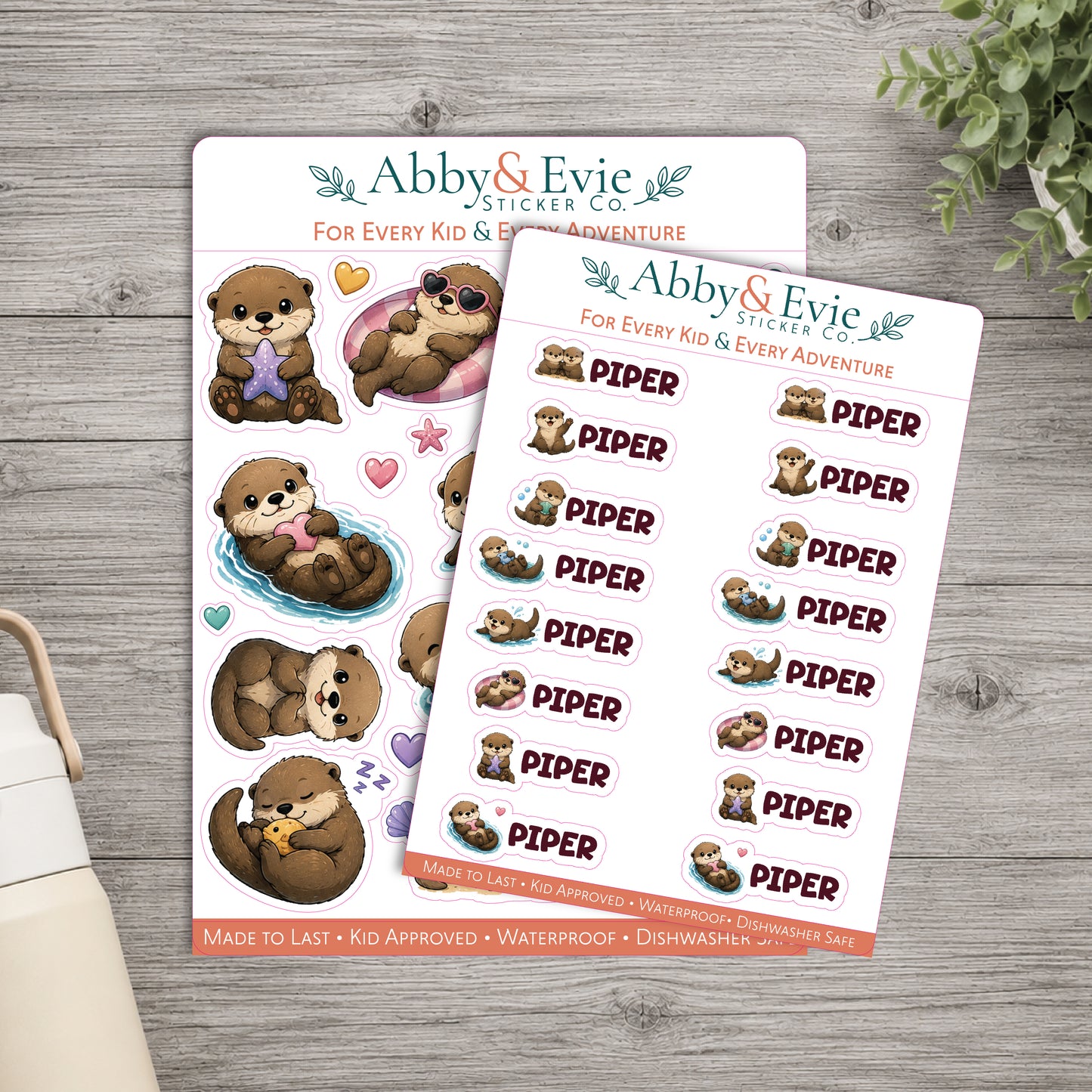 Two sticker sheets from Abby & Evie Sticker Co. featuring cute cartoon sea otters and the word 'PIPER' repeated, styled on a gray wood surface with a small plant and part of a mug visible.