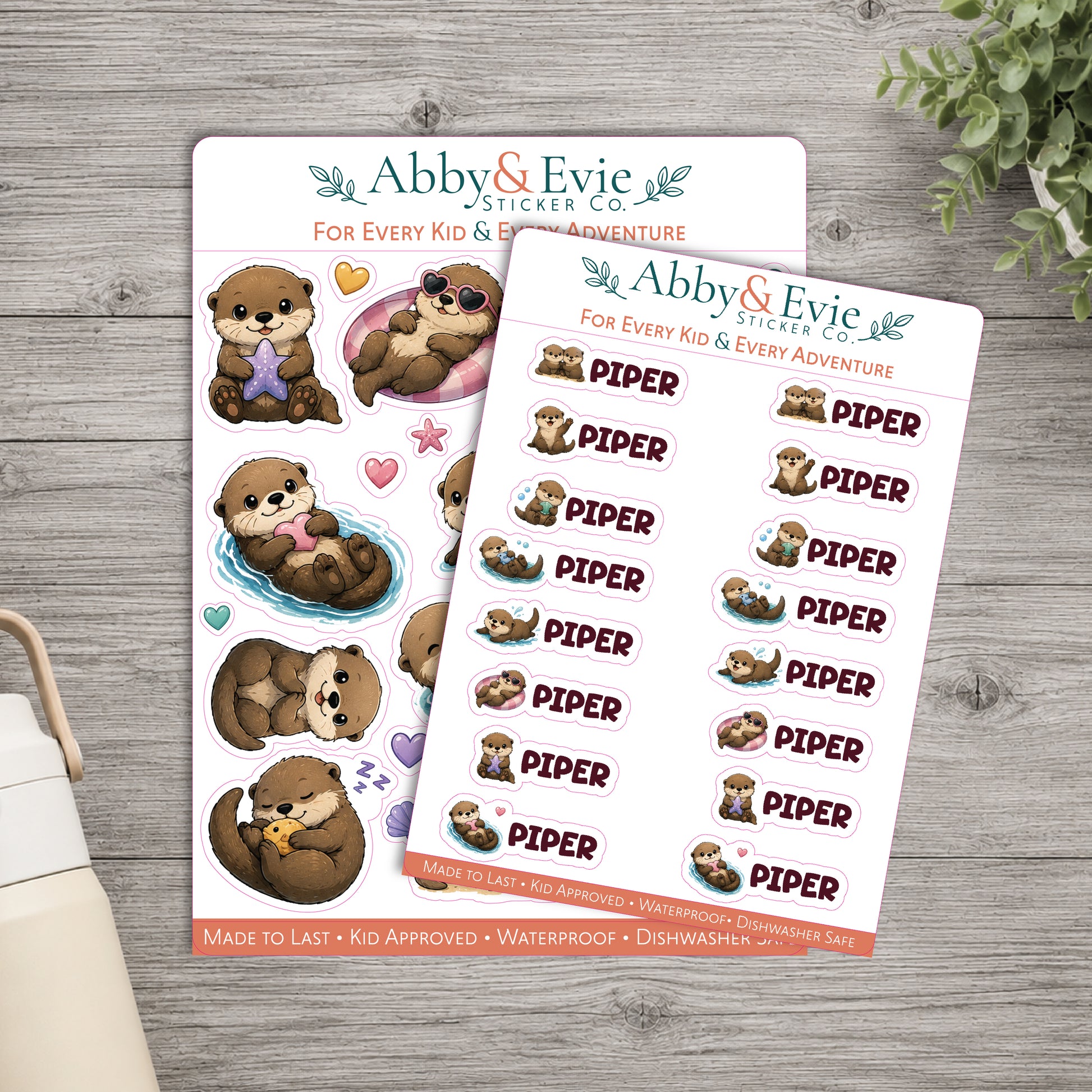 Two sticker sheets from Abby & Evie Sticker Co. featuring cute cartoon sea otters and the word 'PIPER' repeated, styled on a gray wood surface with a small plant and part of a mug visible.