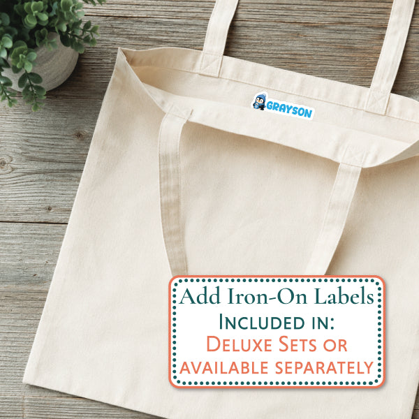 A natural canvas tote bag laid flat on a rustic wood surface, with a small blue and white 'GRAYSON' logo printed inside the neck. A large text overlay at the bottom right reads, 'Add Iron-On Labels INCLUDED IN: DELUXE SETS OR AVAILABLE SEPARATELY.