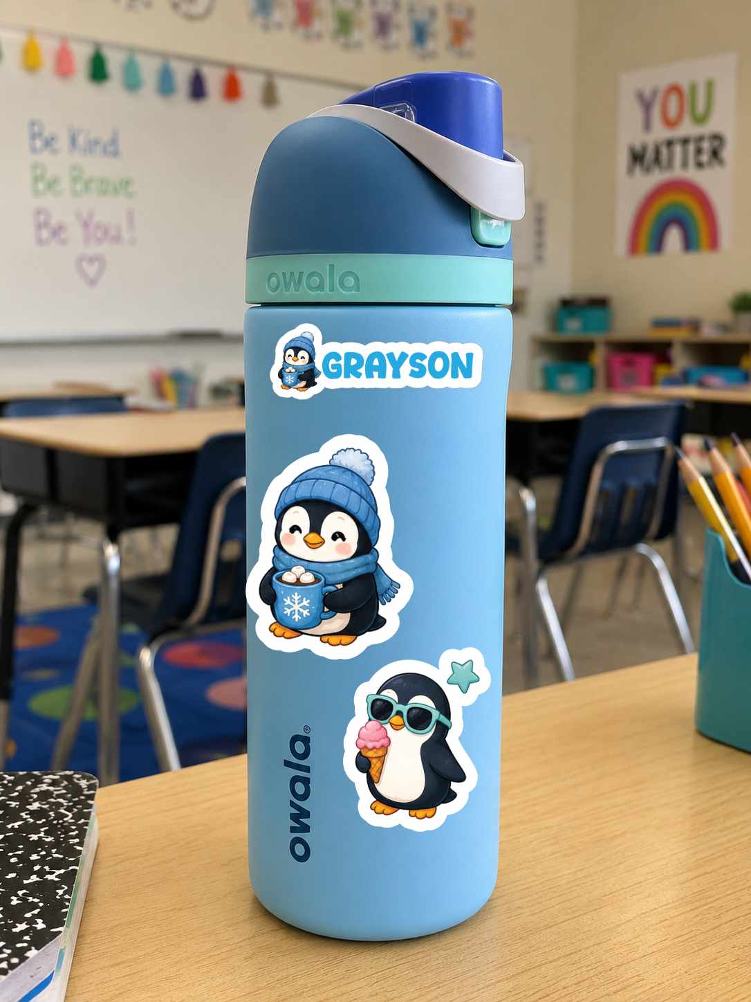 A light blue Owala water bottle with penguin stickers, featuring a custom name decal 'GRAYSON' on the lid, set on a wooden desk in a classroom.