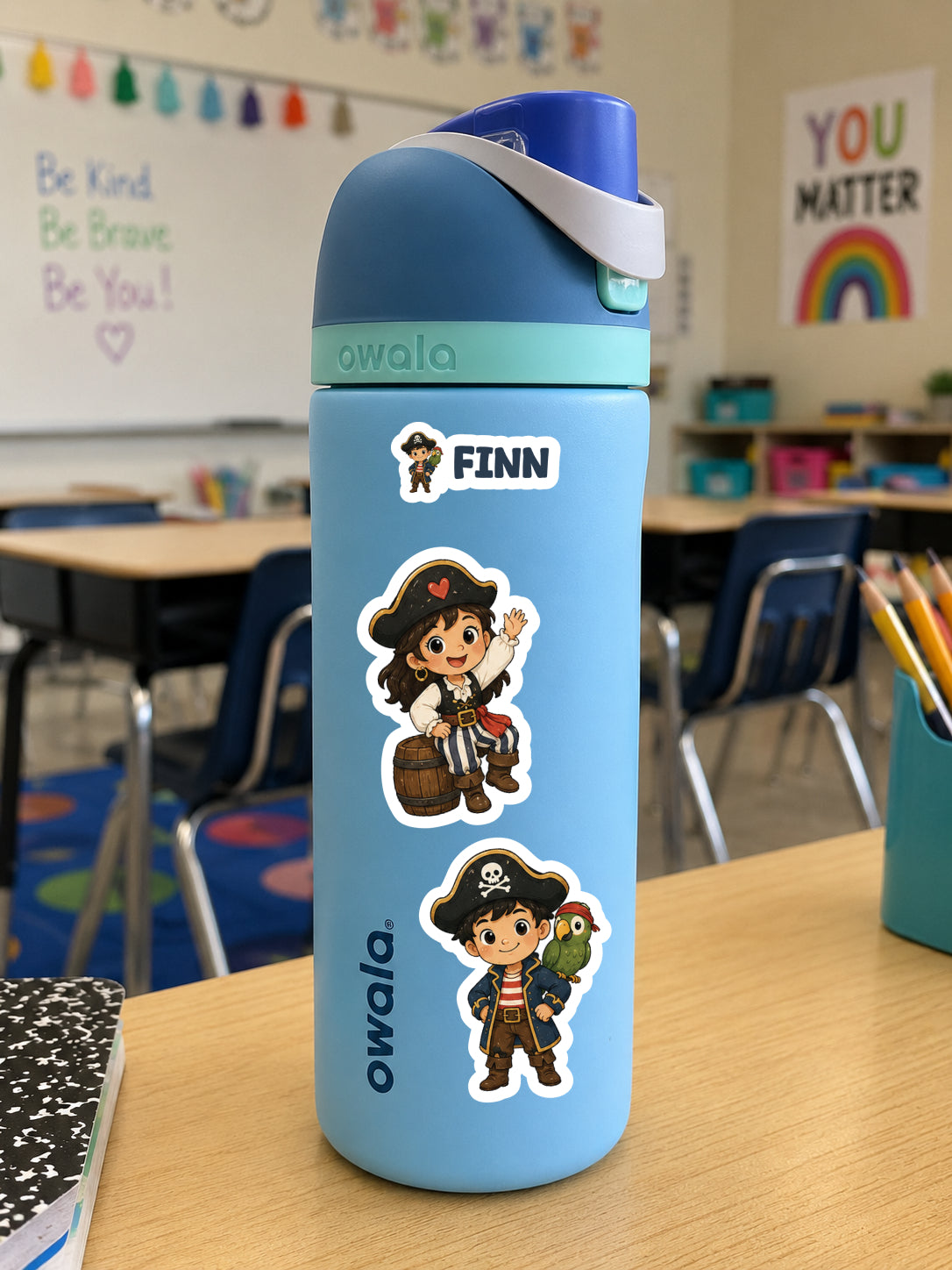 A light blue Owala water bottle with a dark blue lid and white strap, decorated with pirate-themed stickers including the name 'FINN', sitting on a wooden desk in a classroom.