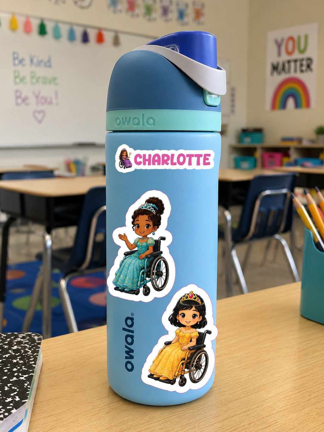 A light blue Owala water bottle with a dark blue lid and a white carrying strap, decorated with two cartoon princess stickers in wheelchairs and a pink 'CHARLOTTE' name sticker, sitting on a wooden desk in a classroom.