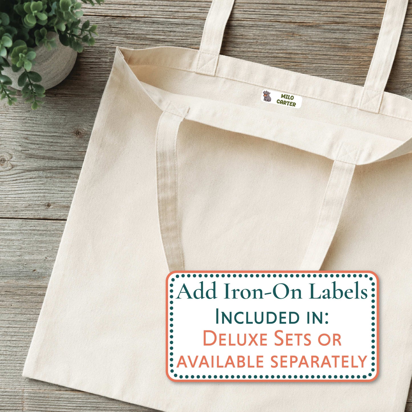 A natural canvas tote bag laid flat on a rustic wood surface, with a small blue and white 'GRAYSON' logo printed inside the neck. A large text overlay at the bottom right reads, 'Add Iron-On Labels INCLUDED IN: DELUXE SETS OR AVAILABLE SEPARATELY.