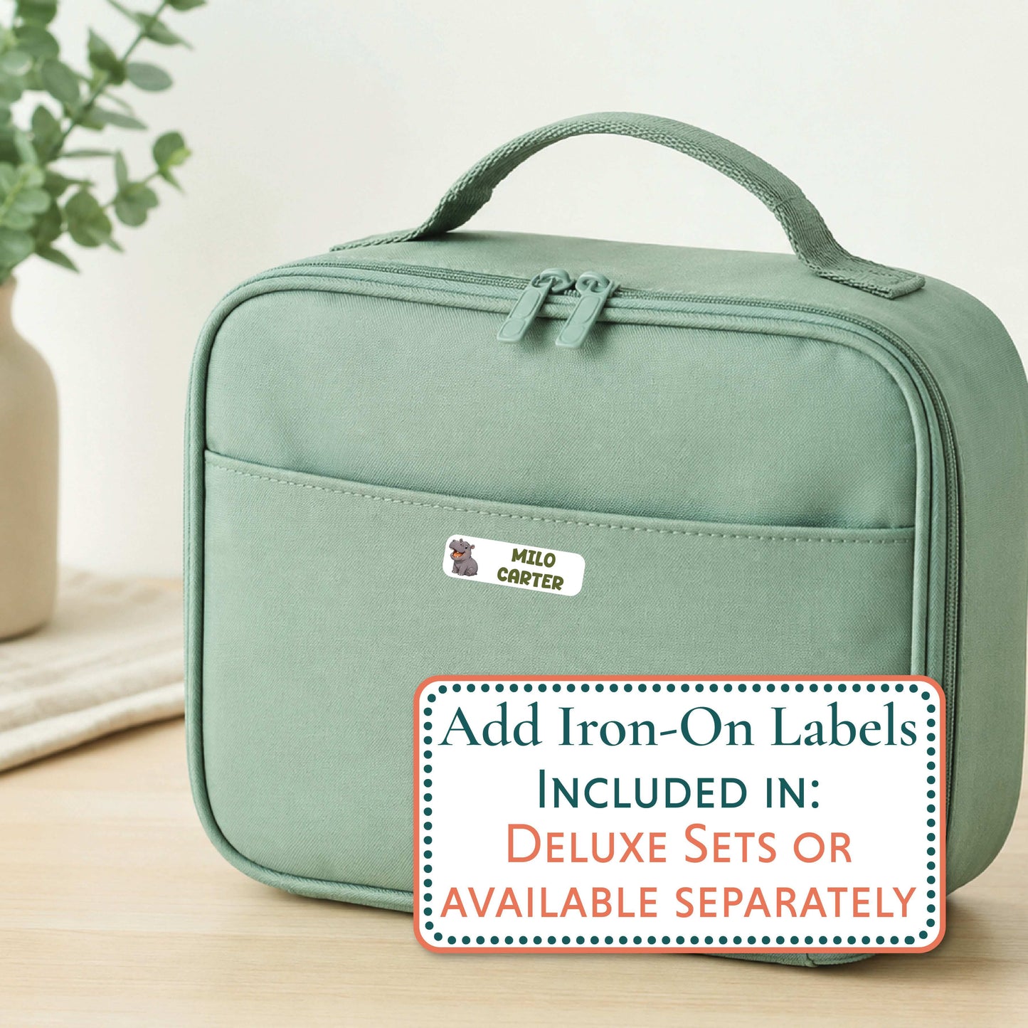 A sage green insulated lunch bag with a top handle and front zip pocket, shown on a light wood surface with a blurred plant in the background. A sticker on the front reads 'MACKENZIE', and a promotional overlay says 'Add Iron-On Labels INCLUDED IN: DELUXE SETS OR AVAILABLE SEPARATELY'.