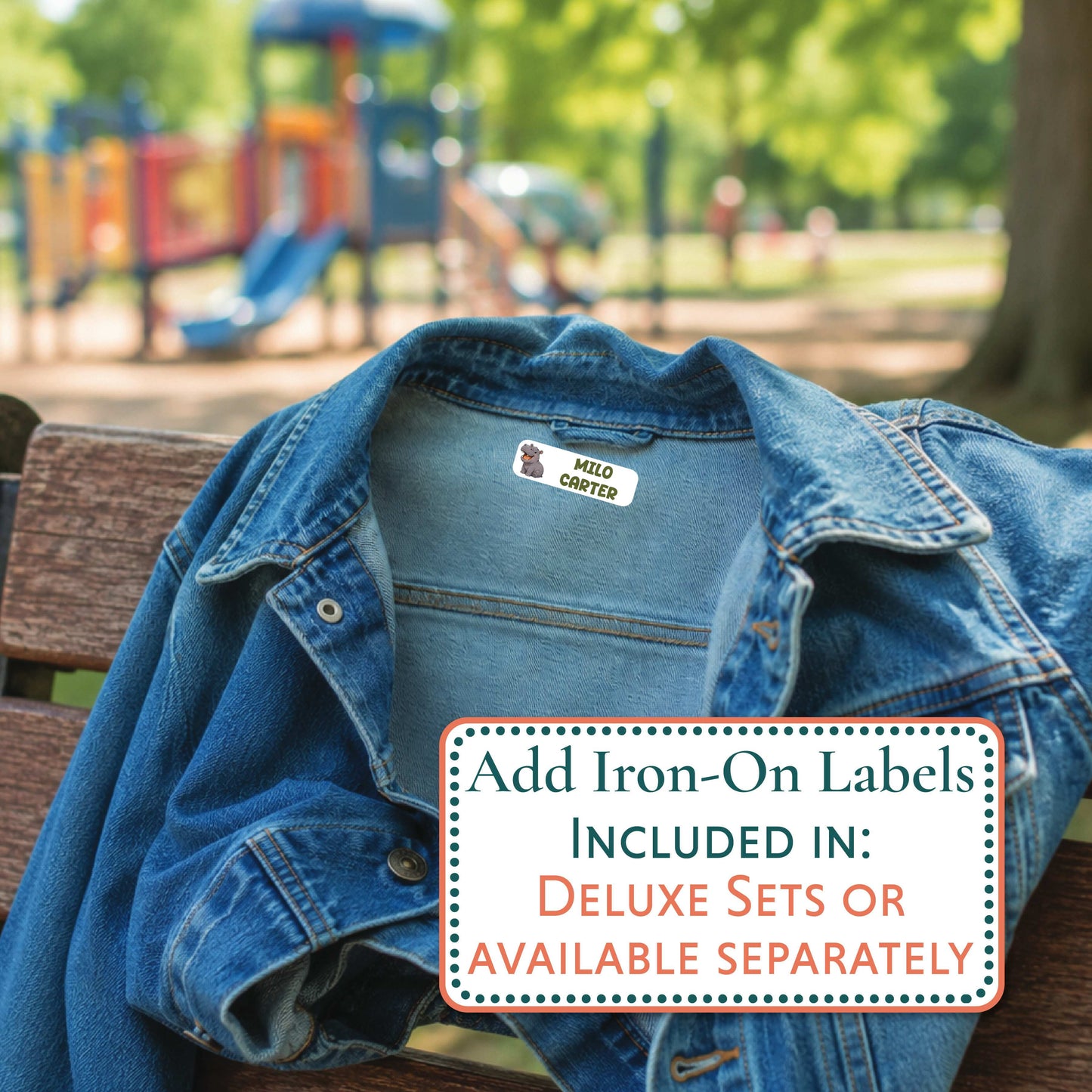 A blue denim jacket draped over a wooden bench in a park, with a blurred playground in the background. A white label on the collar reads CAMERON, and a text box in the foreground says 'Add Iron-On Labels INCLUDED IN: DELUXE SETS OR AVAILABLE SEPARATELY'.