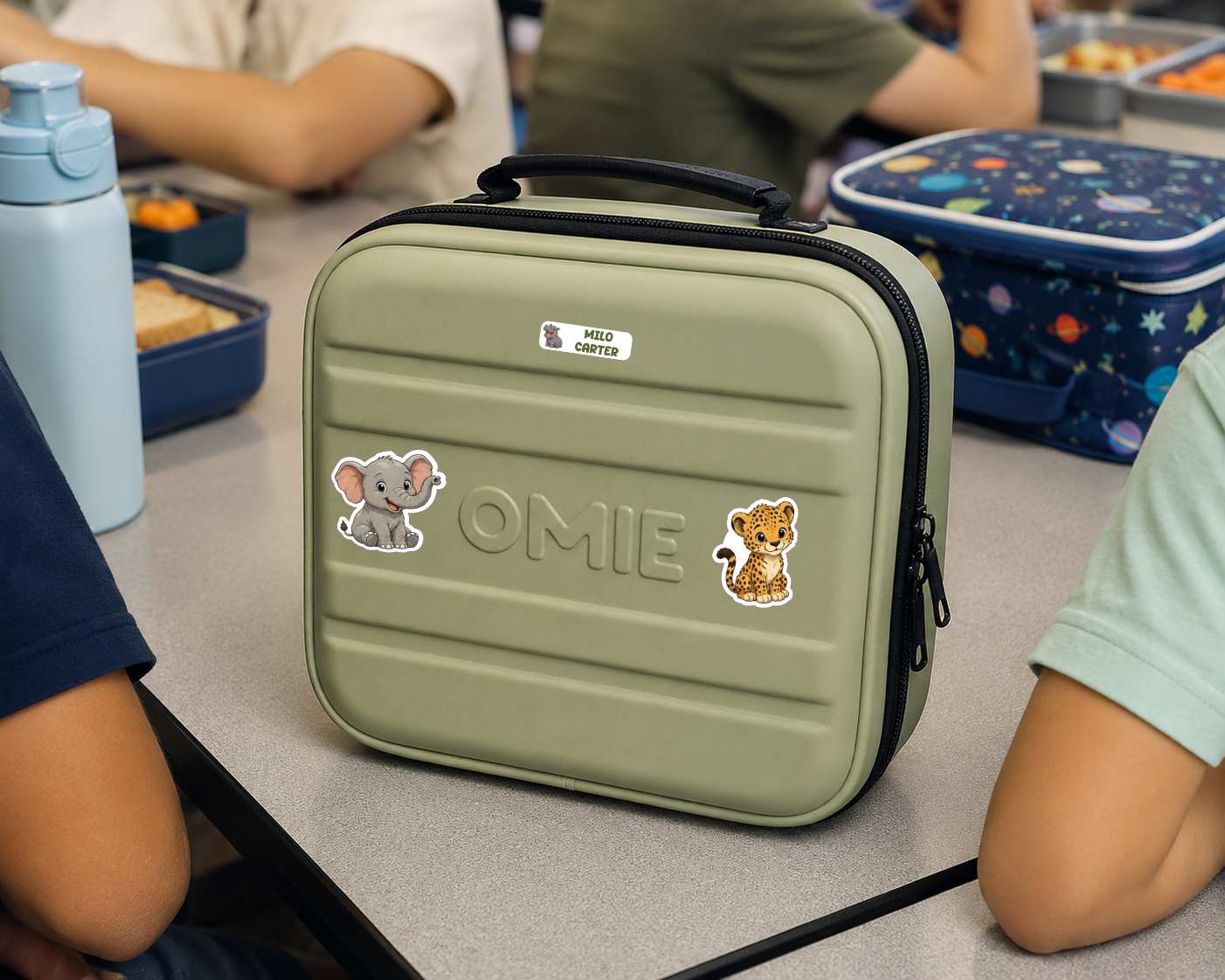 An olive green lunch bag with a black handle and zipper, decorated with stickers and the name 'OMIE' embossed on the front, sitting on a school desk with children around it.