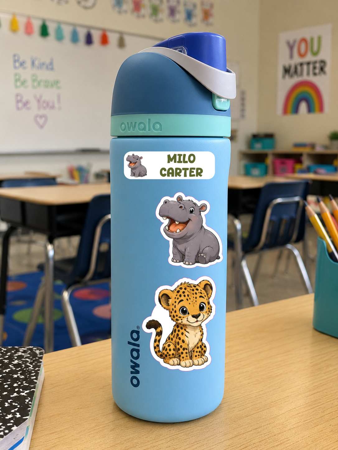 A light blue Owala water bottle with animal stickers sits on a wooden desk in a classroom. The bottle has a custom name label that reads 'MILO CARTER' with a hippo illustration, and other animal stickers below.