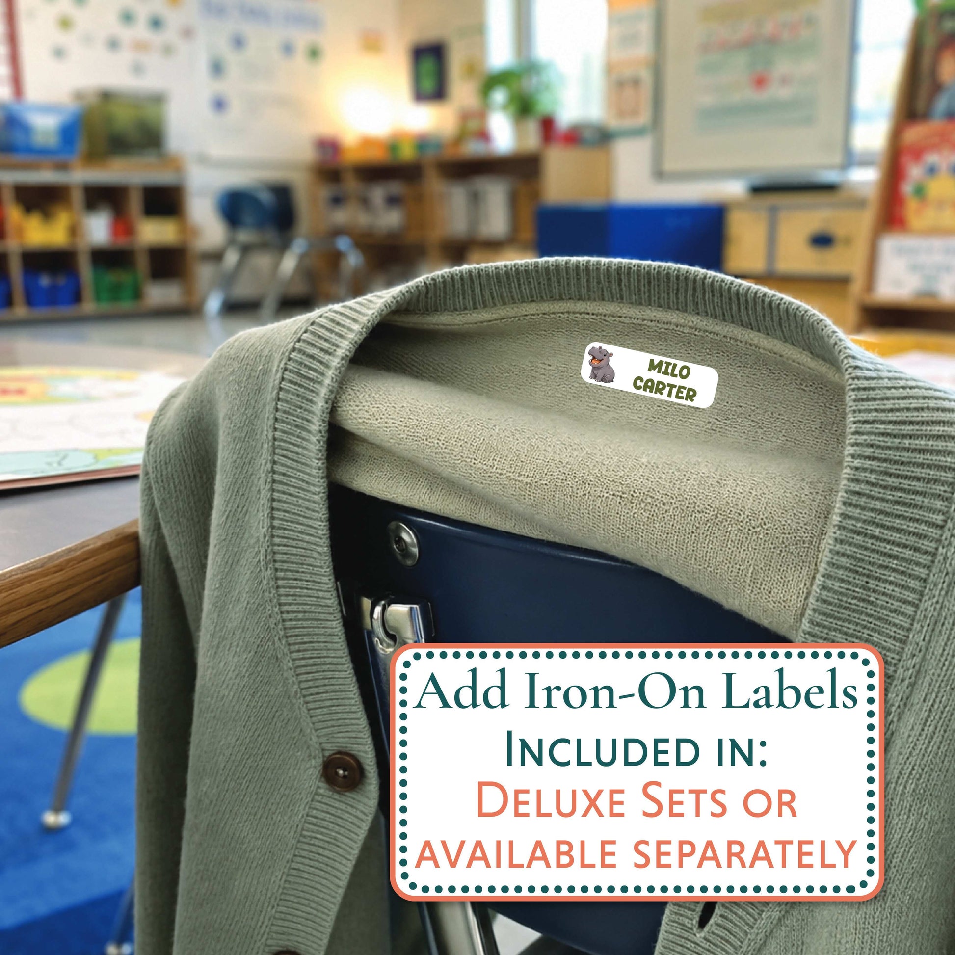 An olive green knit cardigan sweater draped over a dark chair in a blurred classroom setting. A small white and blue 'JORDAN' iron-on label is visible inside the collar, and a text overlay reads 'Add Iron-On Labels INCLUDED IN: DELUXE SETS OR AVAILABLE SEPARATELY'.